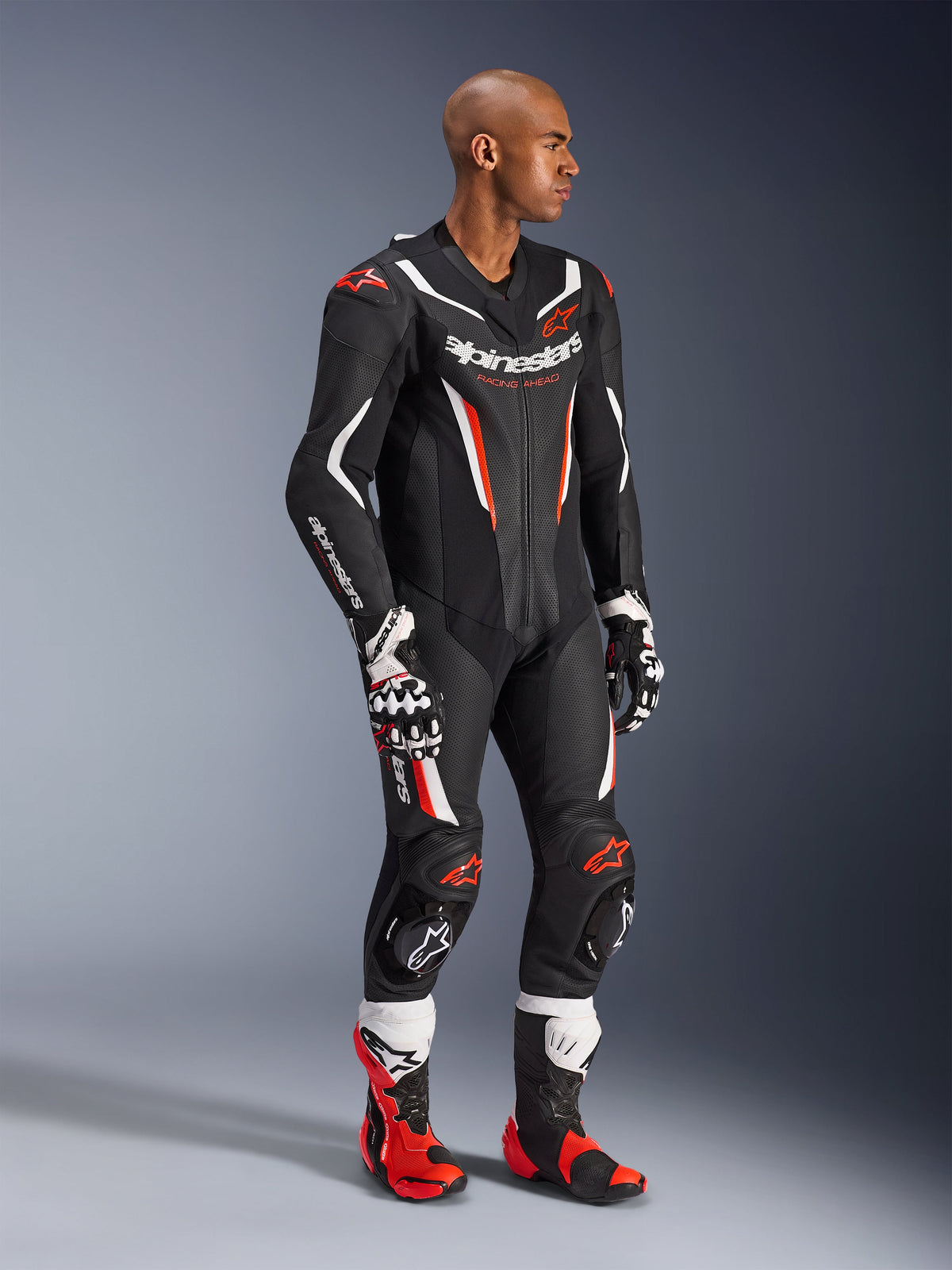 Model iført Alpinestars GP Force V2 1-Piece læderdragt i Black White Red Fluo, højtydende racerudstyr med perforerede læderpaneler og DFS Lite-skjolde, stylet med matchende røde og hvide racerstøvler og handsker, professionel motorsportsæstetik.