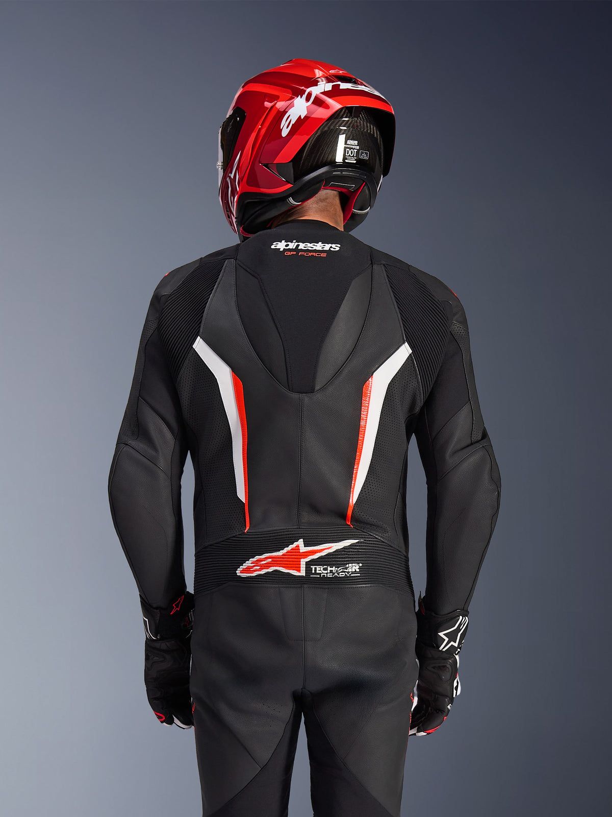 Alpinestars GP Force V2 1-Piece læderdragt, sort, hvid og rød fluo, set bagfra af kører med rød racinghjelm og handsker, med perforerede læderpaneler, aerodynamisk hastighedspukkel med Alpinestars-branding og Tech-Air Ready-logo på den nederste del af ryggen, professionelt motorcykel-racingudstyr.