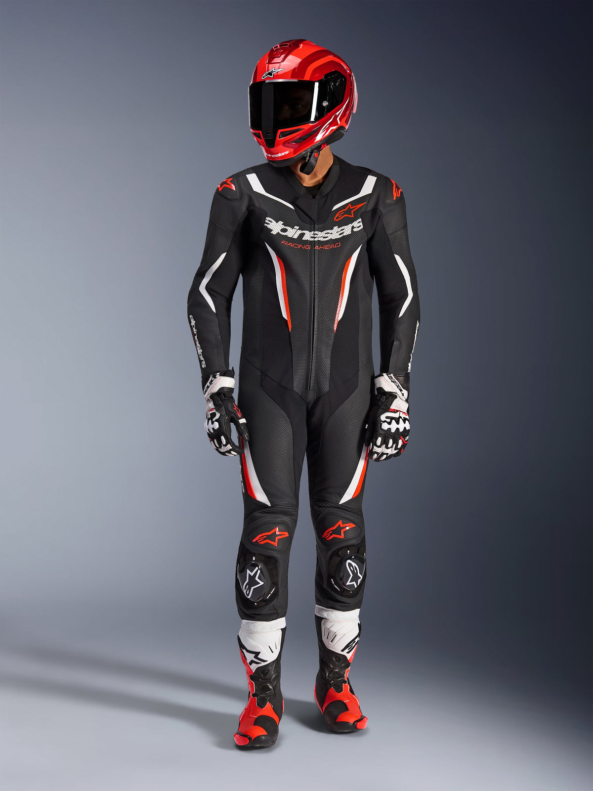 Alpinestars GP Force V2 læderdragt - 1-piece, sort, hvid og rød fluo, båret med en rød racerhjelm, sorte og hvide handsker og røde og sorte støvler, med perforerede læderpaneler og Alpinestars-branding, professionelt motorcykelracerudstyr