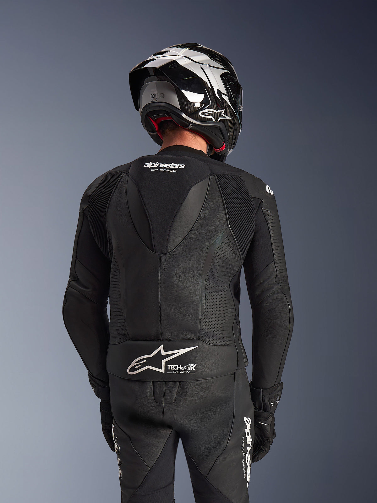 Alpinestars GP Force V2 2-Piece Leather Suit, black, back view of rider showing aerodynamic speed hump, perforated leather panels, and accordion stretch zones on shoulders, with Tech-Air Ready branding on the lower back, worn with matching racing helmet and gloves.