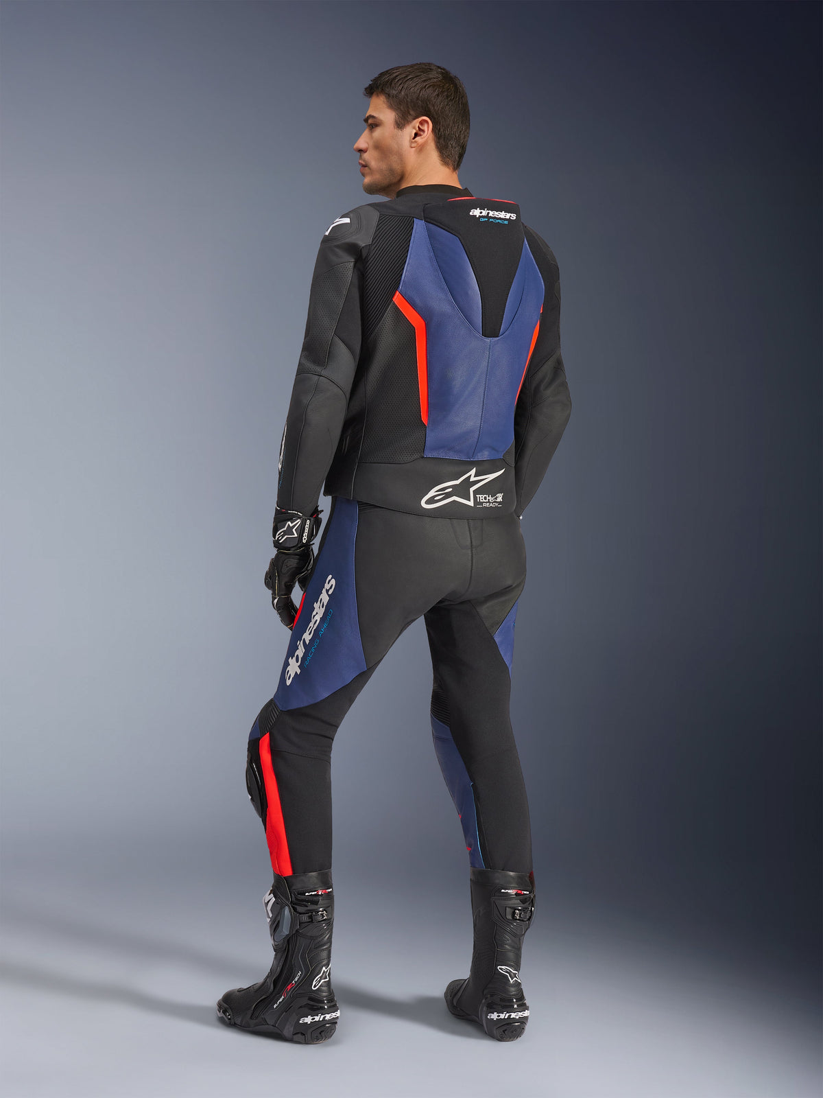 Alpinestars GP Force V2 2-delt læderdragt, sort, blå og rød fluo, profilvisning bagfra på en model, med aerodynamisk pukkel, perforeret læder og Tech-Air Ready-branding, båret med sorte Alpinestars racerstøvler og handsker