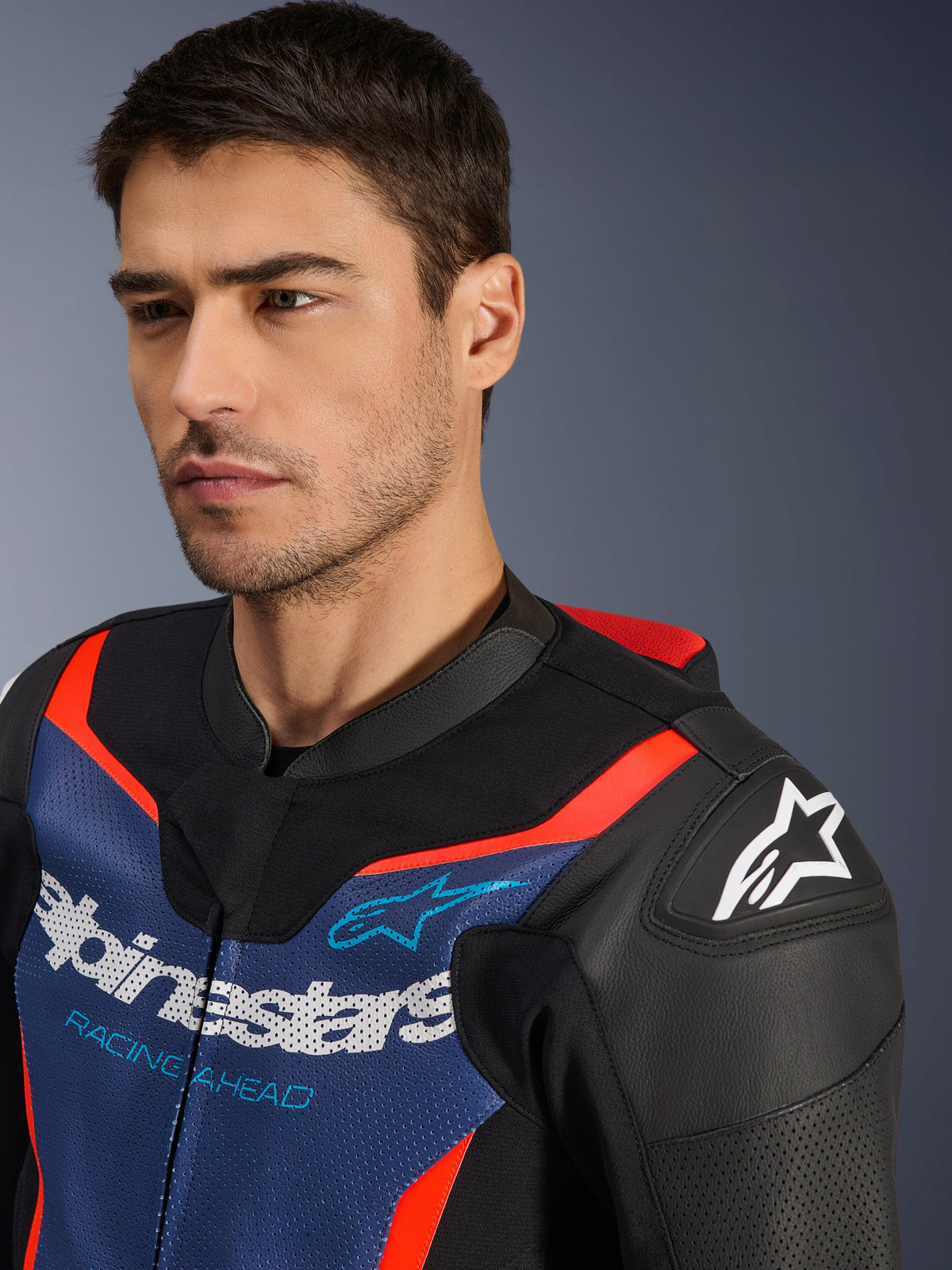 Alpinestars GP Force V2 Læderdragt - 2Piece, sort, blå og rød fluo, nærbillede af en model iført jakken, med perforerede paneler i okselæder, Alpinestars-logo på skulderen og 'Racing Ahead'-branding på brystet, professionelt racingudstyr