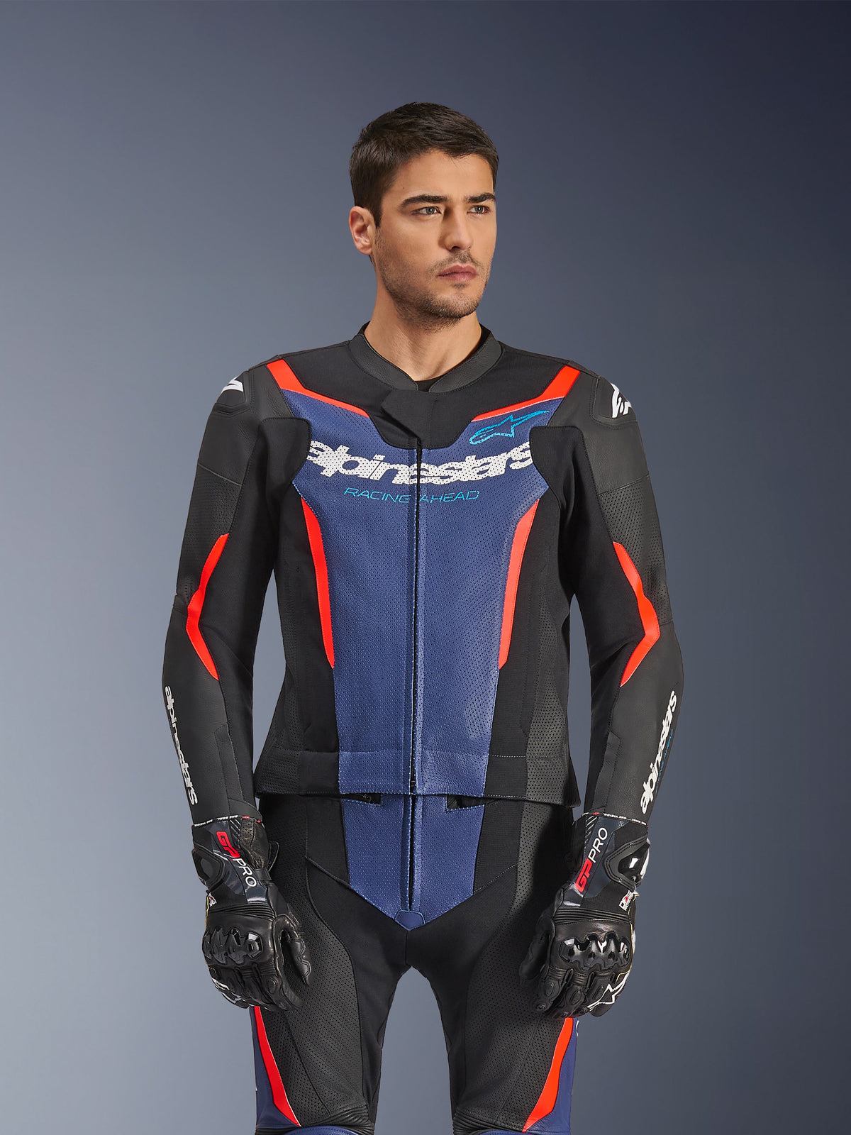 Mandlig model iført Alpinestars GP Force V2 Læderdragt 2-Piece, Sort Blå Rød Fluo, med markant blåt brystpanel og røde detaljer, båret med GP Pro handsker, professionelt motorcykel-racerudstyr i læder med perforeret struktur
