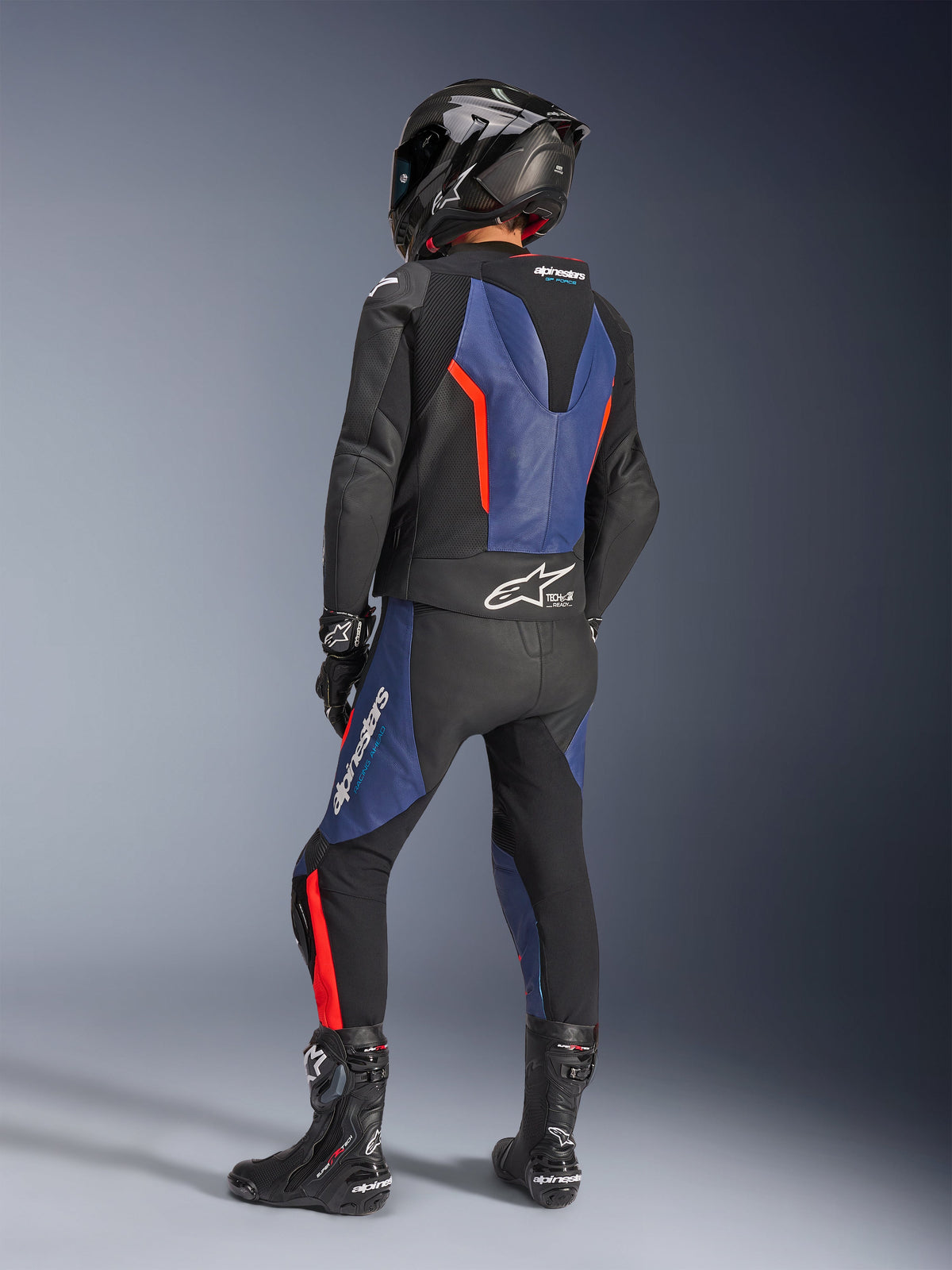 Alpinestars GP Force V2 læderdragt, 2-delt, Sort Blå Rød Fluo, set bagfra af kører i professionelt racerudstyr, med integreret speed hump og Tech-Air Ready branding, blå og rød fluo detaljer på sort læder, kulfiberhjelm og sorte støvler, højtydende motorcykelbeklædning