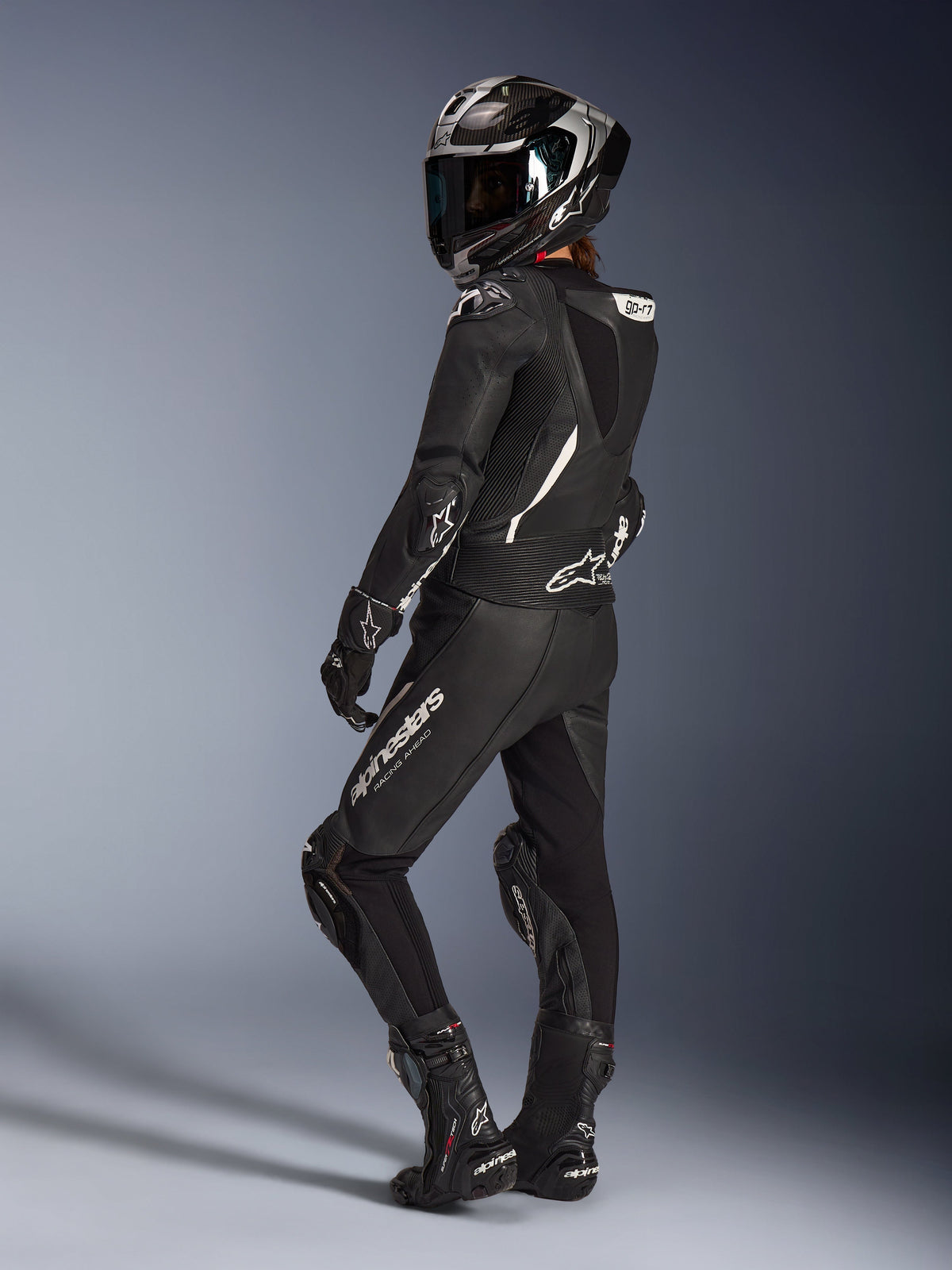 Alpinestars Stella GP-R7 2-delt læderdragt til kvinder, sort, full-face racerhjelm med carbon-finish, matchende handsker og støvler, set bagfra med aerodynamisk pukkel og hvide Alpinestars-logoer, professionelt udstyr til bane- og sportskørsel