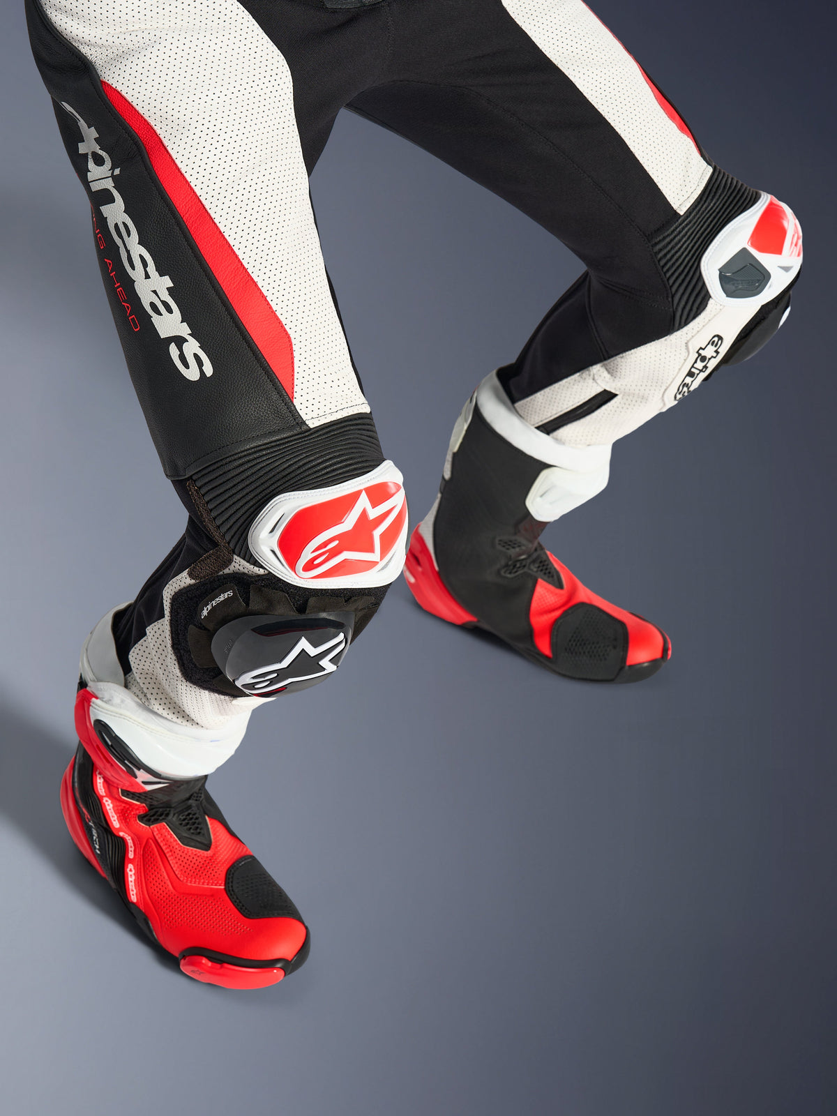 Alpinestars Stella GP-R7 2-delt læderdragt til kvinder, underben og racingstøvler, sort, hvid og rød fluo, med perforeret læder, tydeligt Alpinestars-logo på låret, harmonika-strækpaneler og rød-hvide knæslidere med matchende røde og sorte tekniske støvler.