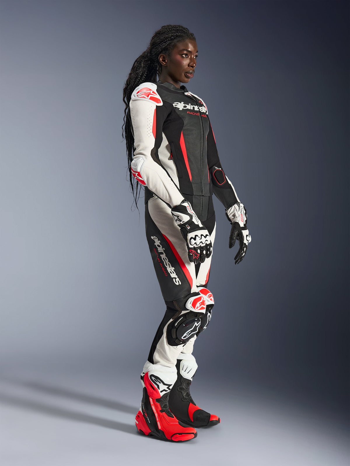 Kvindelig rytter iført Alpinestars Stella GP-R7 2-delt læderdragt i sort, hvid og rød fluo, kombineret med matchende racinghandsker og støvler, med perforerede læderpaneler og beskyttelse på skuldre, albuer og knæ, stående i et studiemiljø.