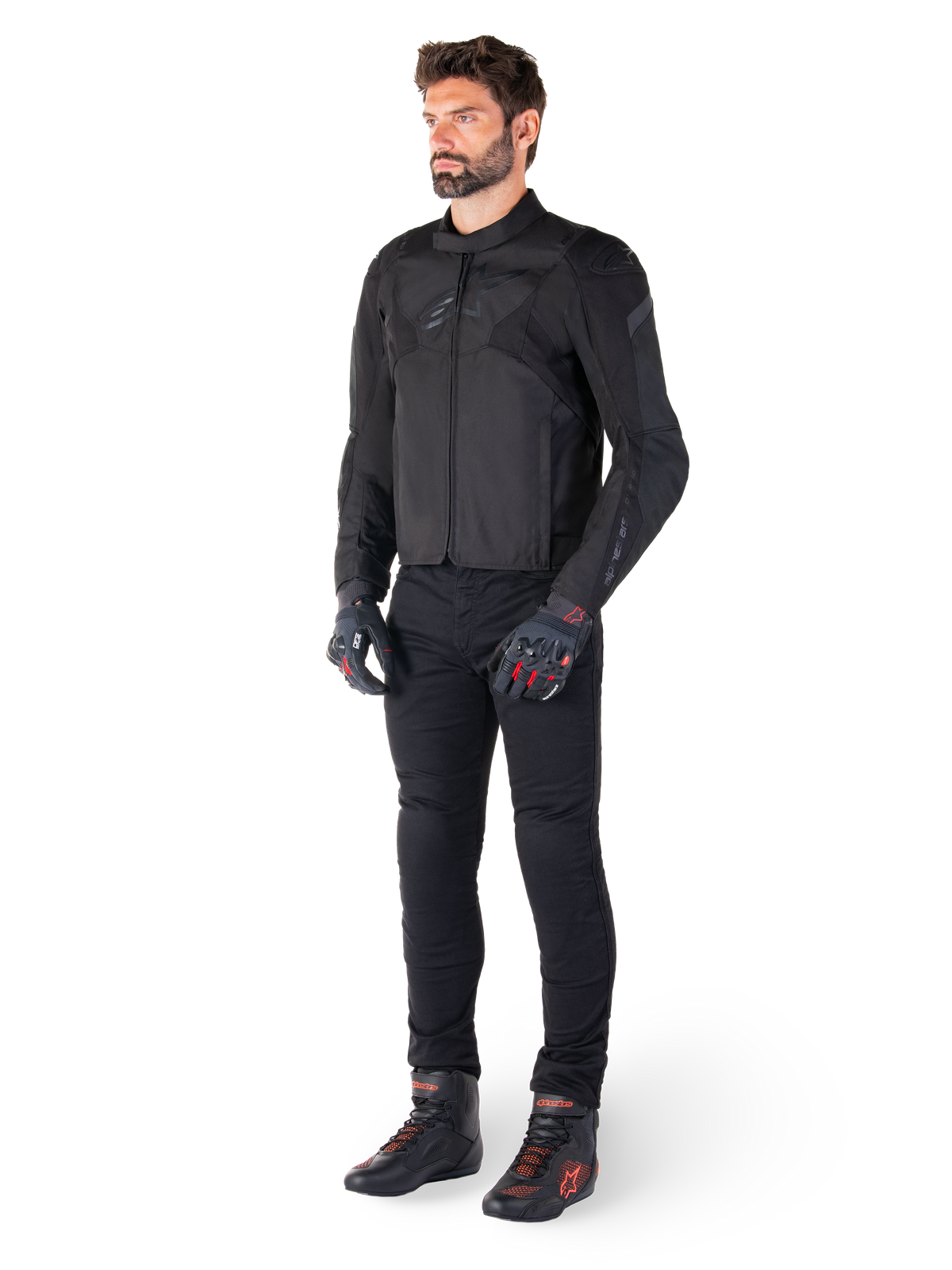 Mandlig model iført Alpinestars T-Jaws V4 vandtæt jakke i sort, kombineret med sorte motorcykelbukser, sorte handsker med røde detaljer og sorte motorcykelsko, strømlinet sportspasform og diskret branding, isoleret på hvid baggrund.