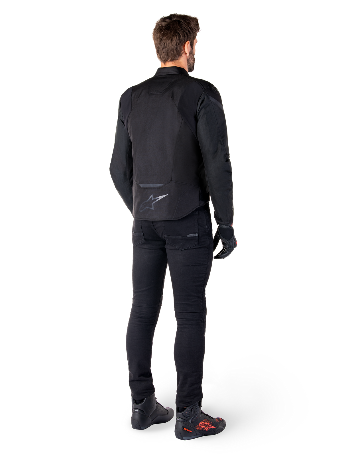 Alpinestars T-Jaws V4 Waterproof Jacket, sort, set bagfra på model iført sorte kørebukser og støvler, der viser sportstekstilkonstruktionen og Alpinestars-logoet på den nederste del af ryggen.