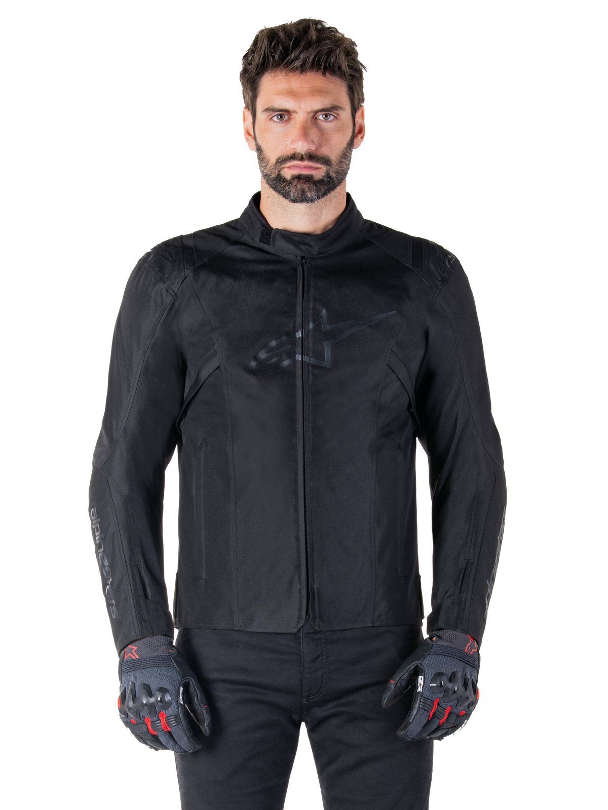 Alpinestars T-SPS V2 Waterproof Jacket, sort motorcykel-tekstiljakke, båret af kører med sorte og røde handsker, med høj krave, lynlås foran og diskret logo på brystet, vandtæt beskyttelse til sport- og bykørsel