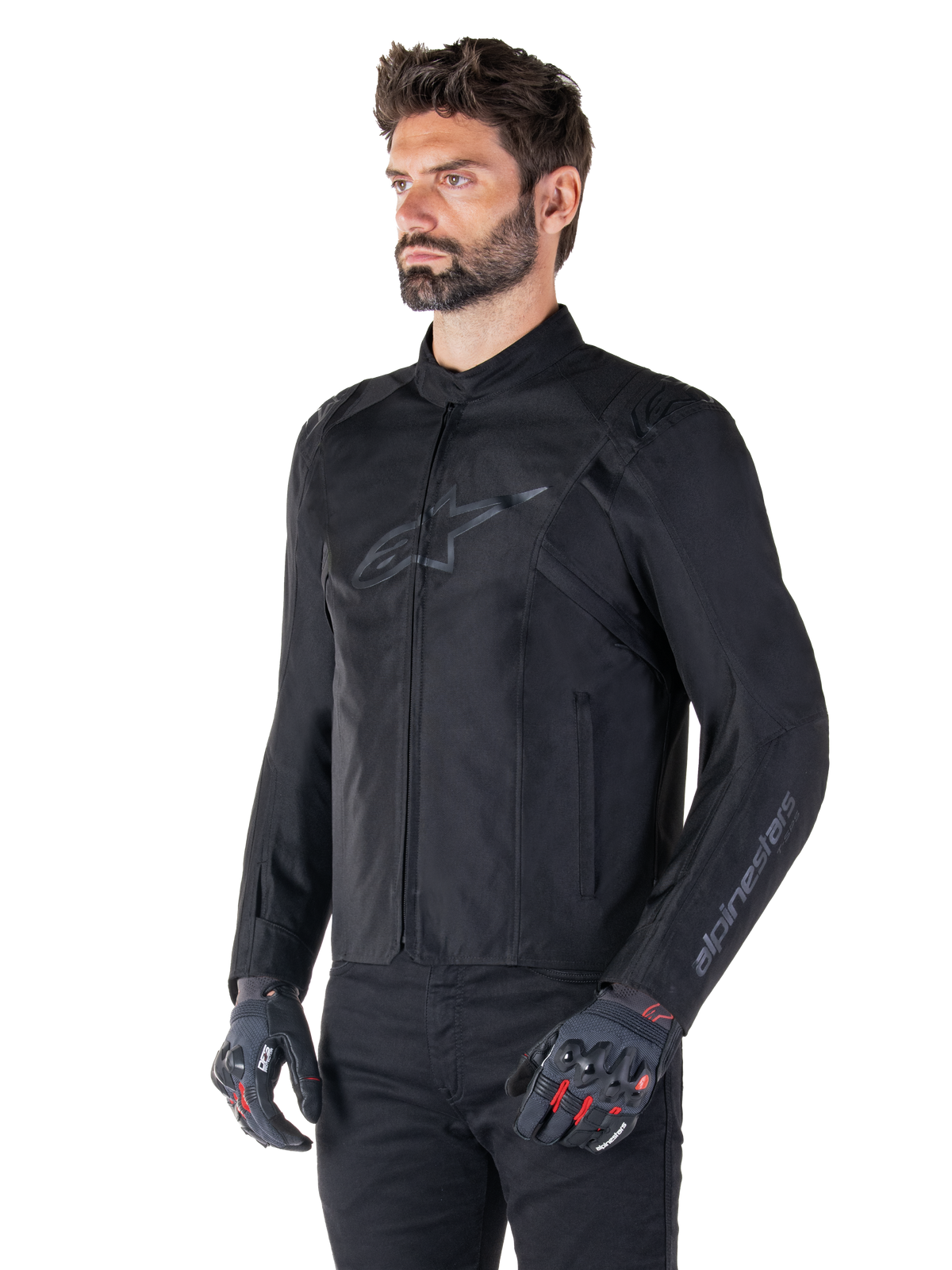Model iført Alpinestars T-SPS V2 vandtæt jakke, sort, kombineret med sorte bukser og grå motorcykelhandsker med røde detaljer, diskret sort brystlogo og branding på ærmet, tekstiljakke i sportssnit til landevejskørsel.