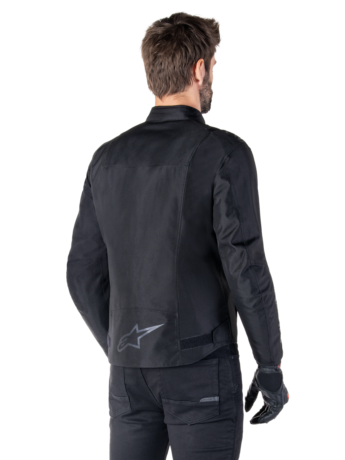Alpinestars T-SPS V2 Vandtæt Jakke, sort, set bagfra på en mandlig model, med et reflekterende stjerne-logo nederst på ryggen, forlænget rygprofil og taljejusteringsstropper, kombineret med sorte kørebukser og handsker