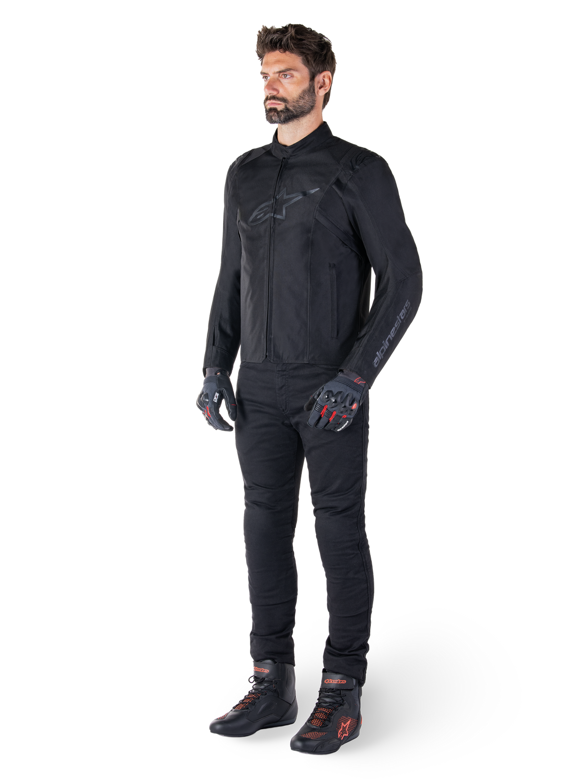 Alpinestars T-SPS V2 vandtæt jakke, sort, båret af en mandlig model med matchende sorte bukser, sorte handsker med røde detaljer og sorte køresko, komplet motorcykeludstyr til by- og sportskørsel, ren hvid baggrund