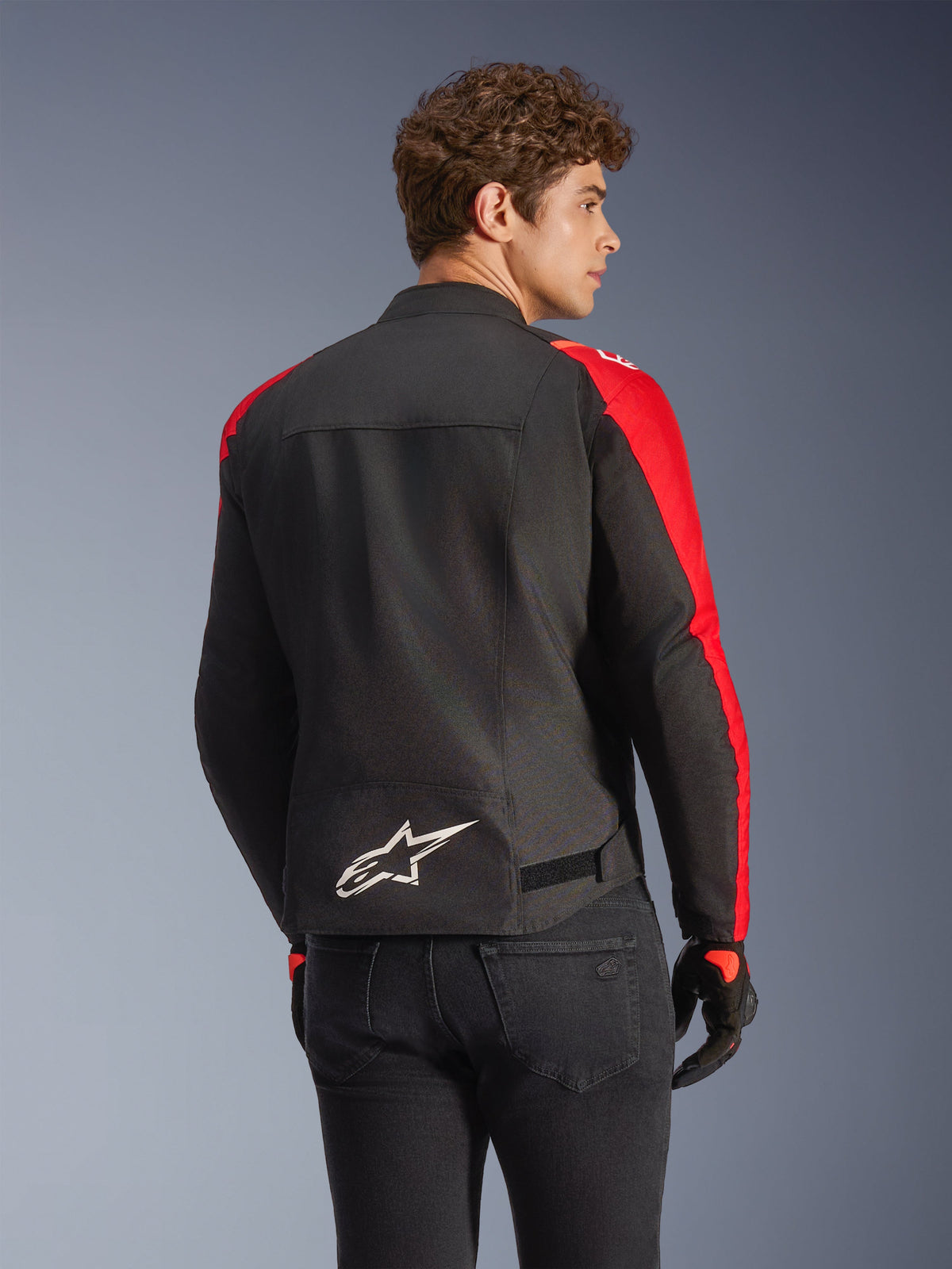 Alpinestars T-SPS V2 vandtæt jakke i sort og rød, set bagfra på en model, med et hvidt Alpinestars-logo nederst på ryggen, stærke røde ærmepaneler og en sport-cut silhuet, båret med sorte handsker og jeans.
