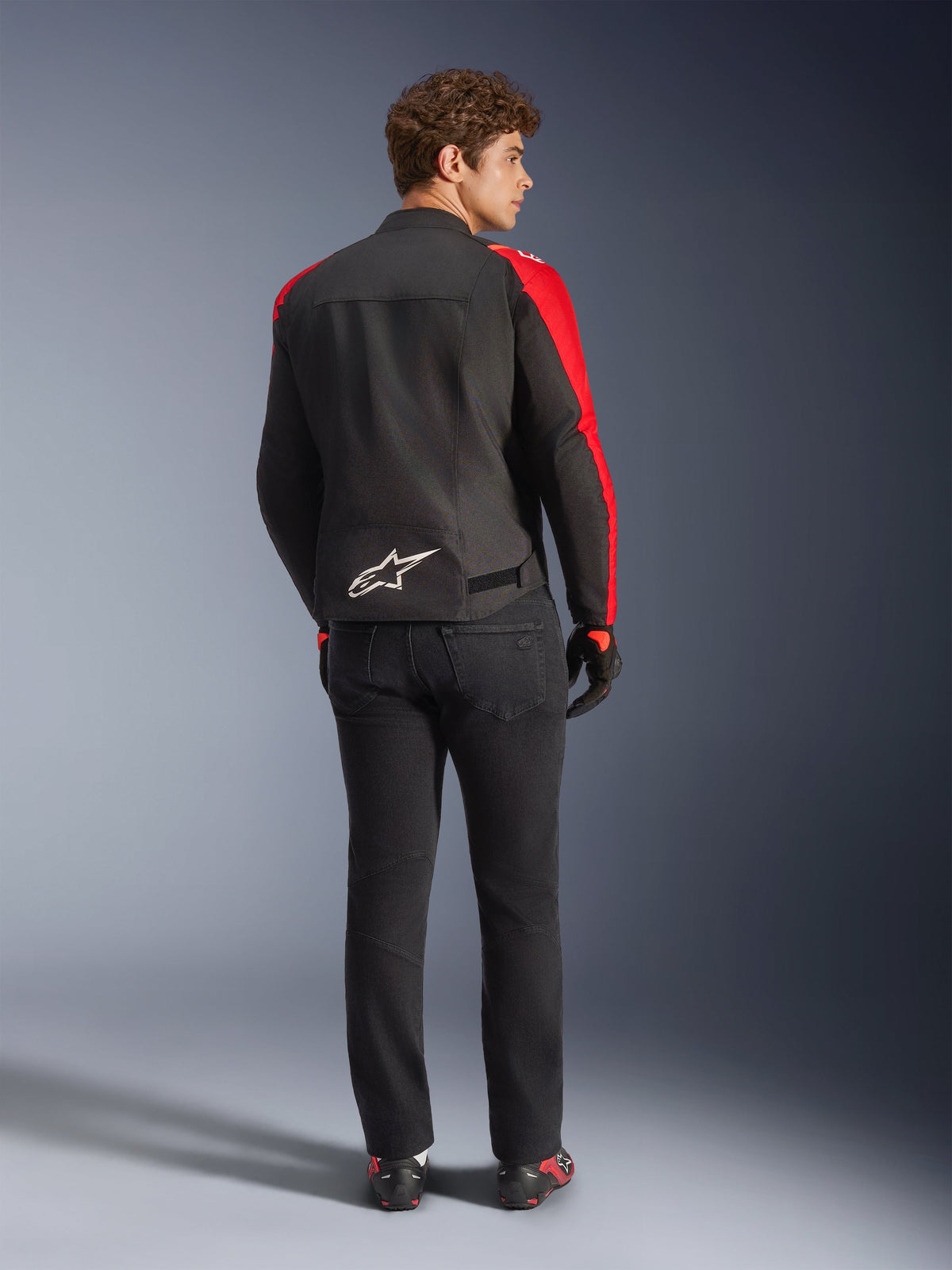 Alpinestars T-SPS V2 Vandtæt Jakke, Sort Rød Rød Fluo, set bagfra på mandlig model, hvidt Alpinestars-logo nederst på ryggen, båret med sorte motorcykelbukser og handsker, sporty vandtæt tekstiljakke