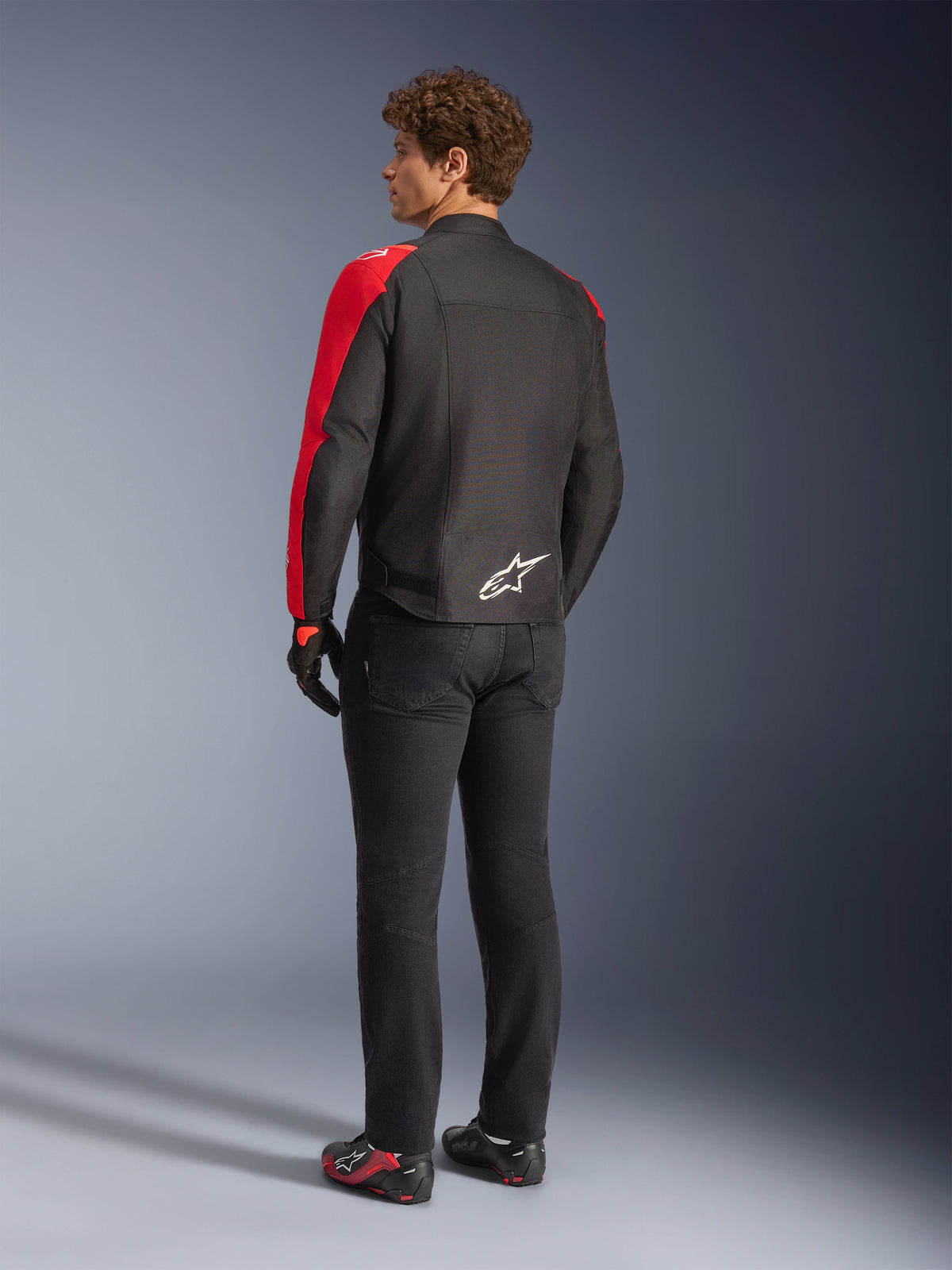 Alpinestars T-SPS V2 vandtæt jakke i sort og rød fluo, set bagfra på model, båret med sorte motorcykelbukser og køresko, hvidt Alpinestars-logo på lænden, tekstiljakke i sports-snit
