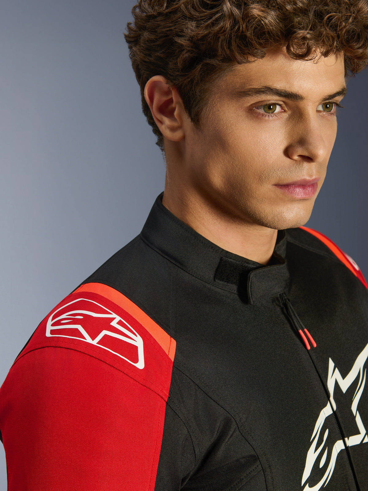 Alpinestars T-SPS V2 vandtæt jakke, motorcykeltekstiljakke, sort rød og rød fluo, nærbillede af model, der bærer jakken, med fremtrædende hvide Alpinestars-logoer på skulder og bryst, lavprofilskrave og atletisk sportspasform.