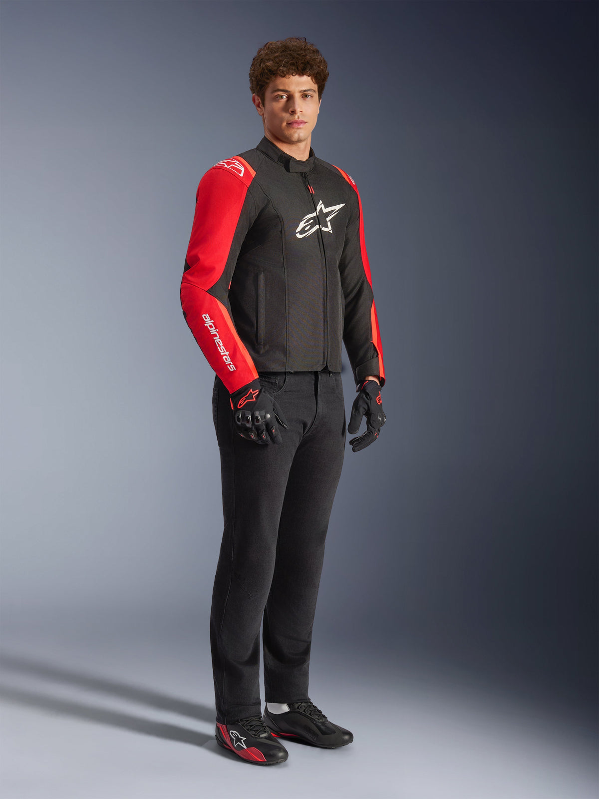 Mandlig model iført Alpinestars T-SPS V2 vandtæt jakke i sort og rød fluo, med markante røde ærmer med hvid branding og et stort hvidt logo på brystet, kombineret med sorte motorcykelbukser, motorcykelhandsker og lavprofil køre-sko, studiebillede i fuld figur.
