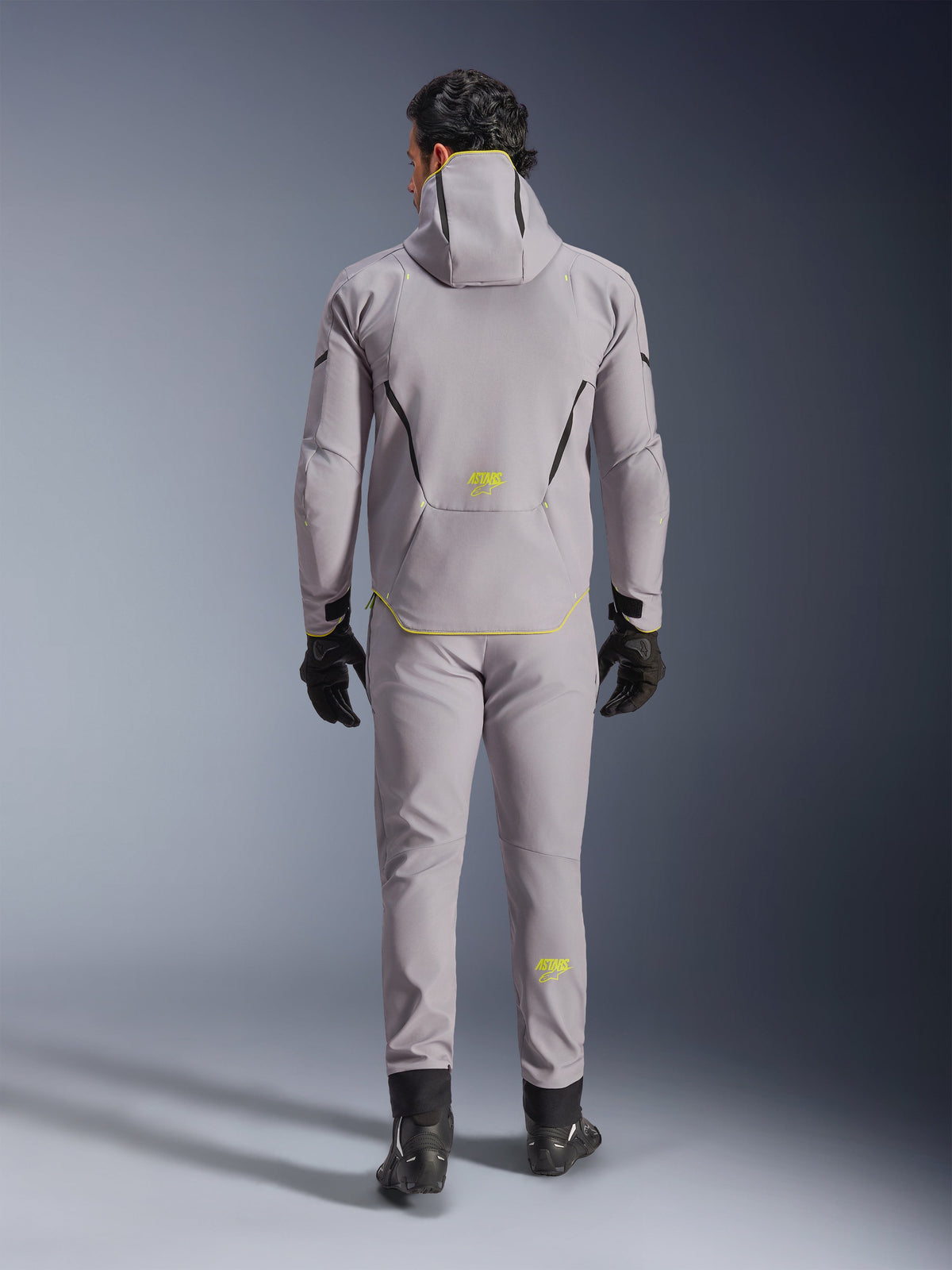 Alpinestars Aeroshell Waterproof Jacket in Mid Gray Yellow Fluo, full back view, worn with matching gray pants, black gloves, and black riding shoes. Features a hooded softshell design with yellow fluo 'ASTARS' branding and accents, engineered for modern motorcycle road performance.