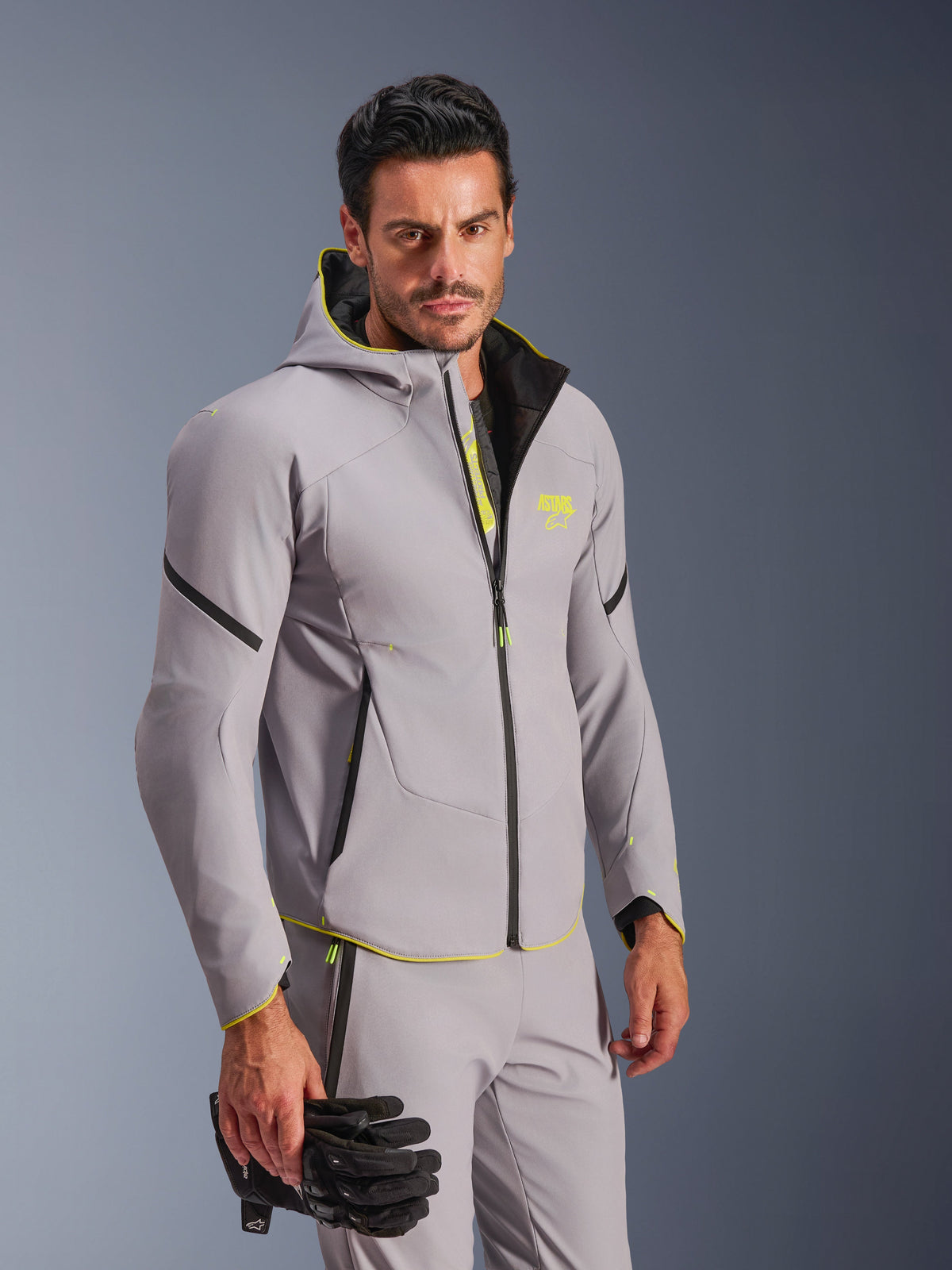 Alpinestars Aeroshell Waterproof Jacket in Mid Gray and Yellow Fluo, worn by a man with matching gray pants and black motorcycle gloves, featuring a hooded softshell design with neon yellow accents and chest logo, modern sports motorcycle gear.