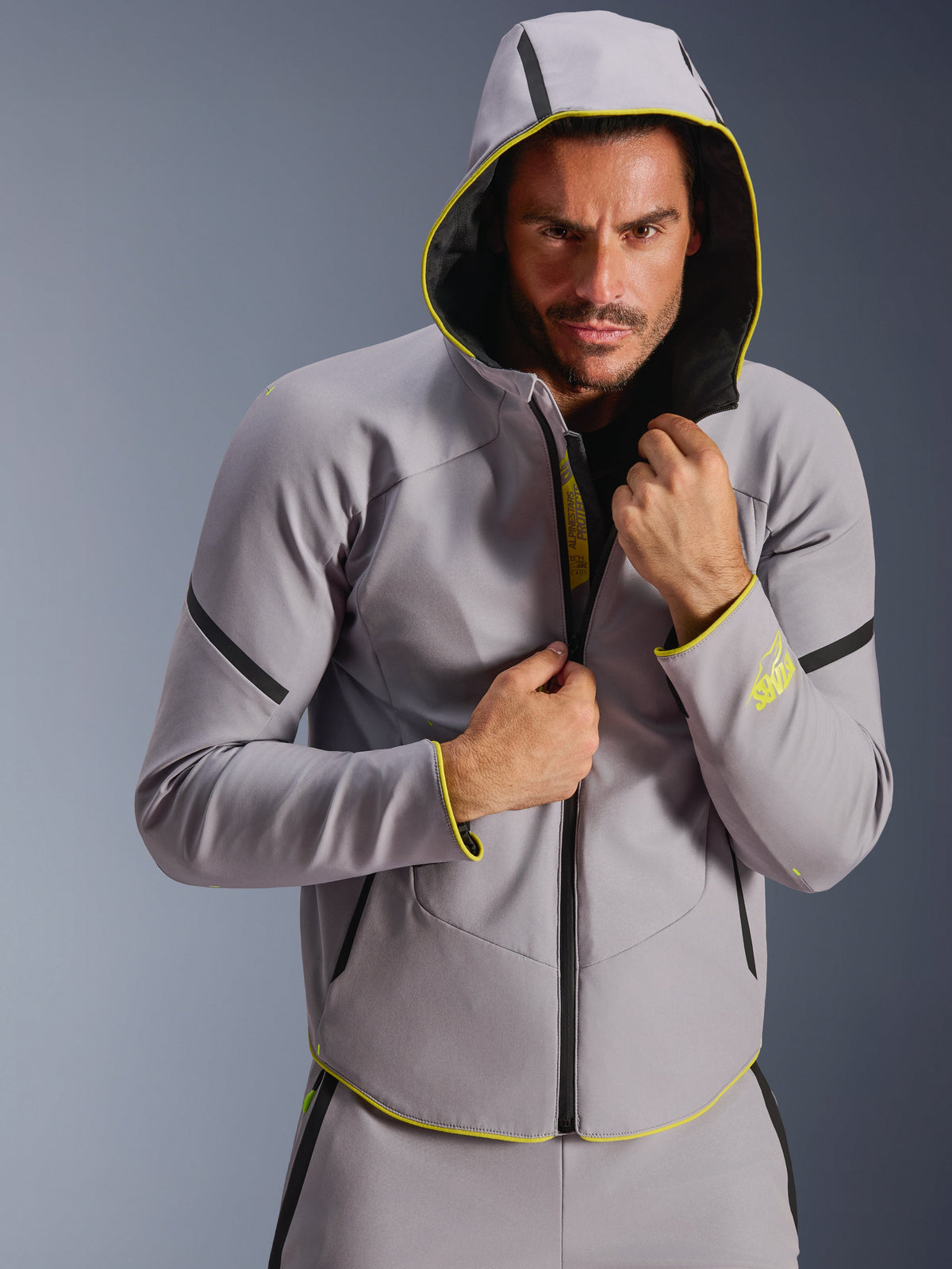 Alpinestars Aeroshell Waterproof Jacket, Mid Gray and Yellow Fluo, sports softshell jacket with a hoodie, worn by a male model with the hood up, featuring black waterproof zippers and fluorescent yellow accents on the cuffs and hem, modern motorsport-inspired design