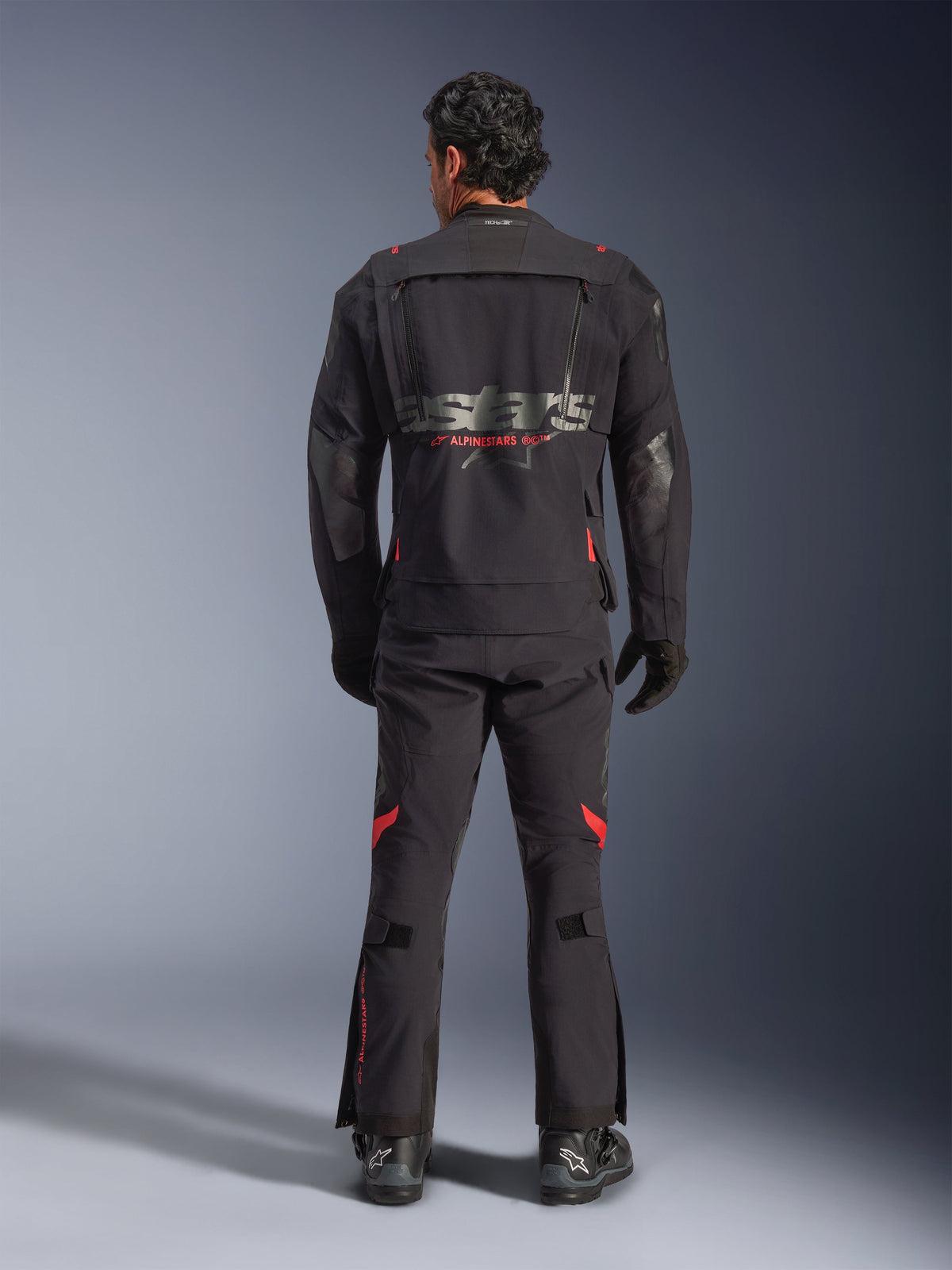 En rytter set bagfra iført Alpinestars Halo Pro Drystar® XF lamineret jakke i Black Dark Shadow, med et stort gråt astars-logo og rød Alpinestars-branding, modulære ventilationspaneler på ryggen, kombineret med matchende adventure-bukser og sorte støvler.