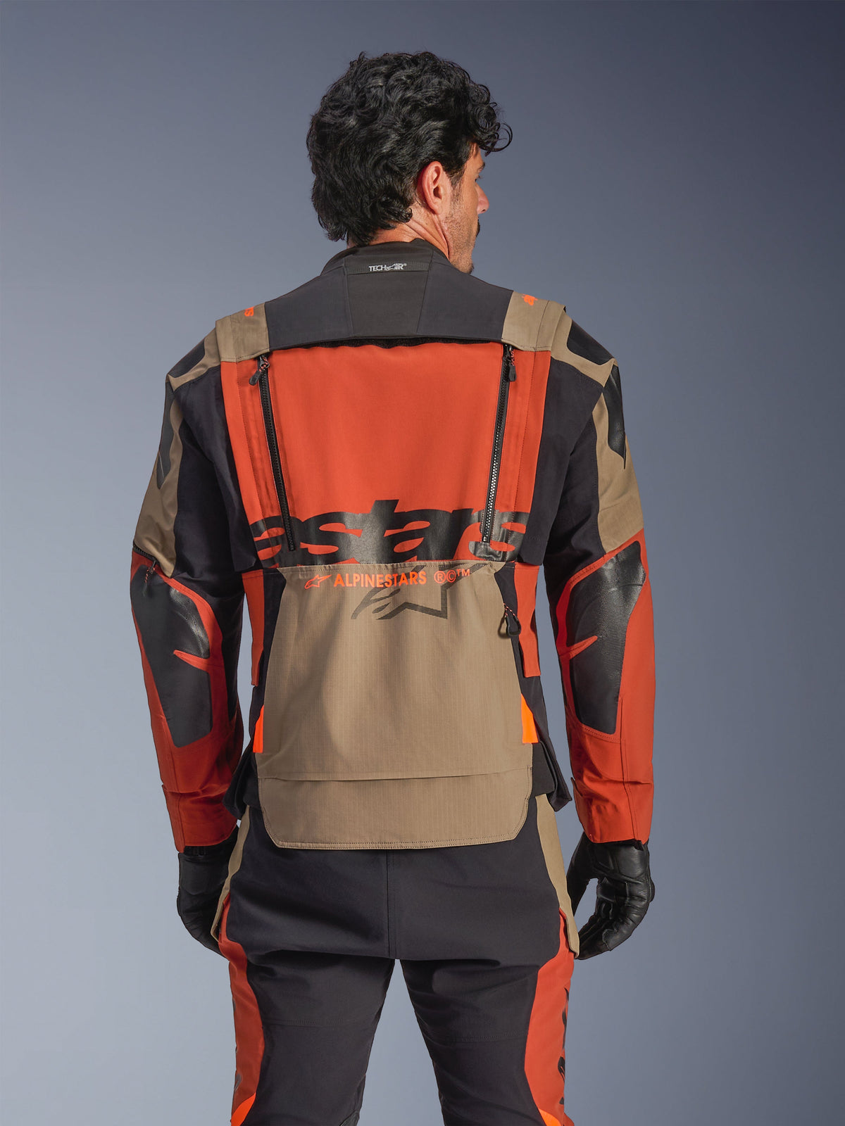 Alpinestars Halo Pro Drystar® XF Laminated Jacket, Walnut Rust Black colorway, rear view, featuring a large orange back panel with black ASTARS branding, walnut lower storage pocket, and Tech-Air® Ready collar, worn with matching adventure riding pants and black gloves