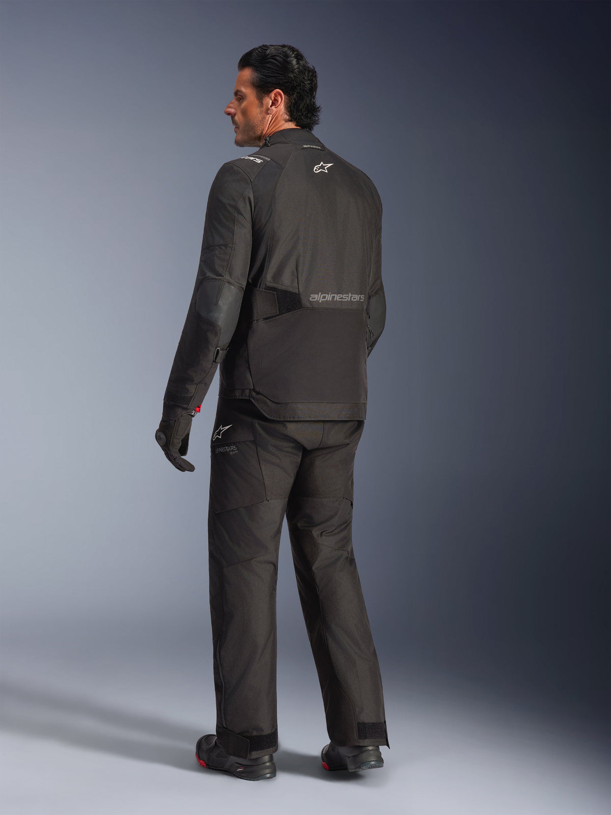Alpinestars Andes V4 Drystar® jakke, sort, rygbillede af kører i komplet motorcykeludstyr, tekstil adventure-jakke med Alpinestars-branding på ryggen, båret med matchende sorte bukser og støvler, all-weather køreudstyr til vej og adventure
