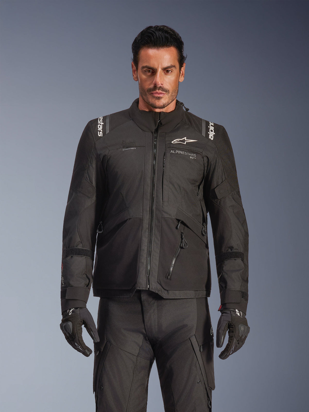 Mandlig model iført Alpinestars Andes V4 Drystar® jakke i sort, adventure touring motorcykeljakke, kombineret med matchende sorte kørebukser og handsker, udstyret med vandtætte lynlåse og hvid Alpinestars-branding på brystet og skuldrene, studiooptagelse af køreudstyr til al slags vejr.