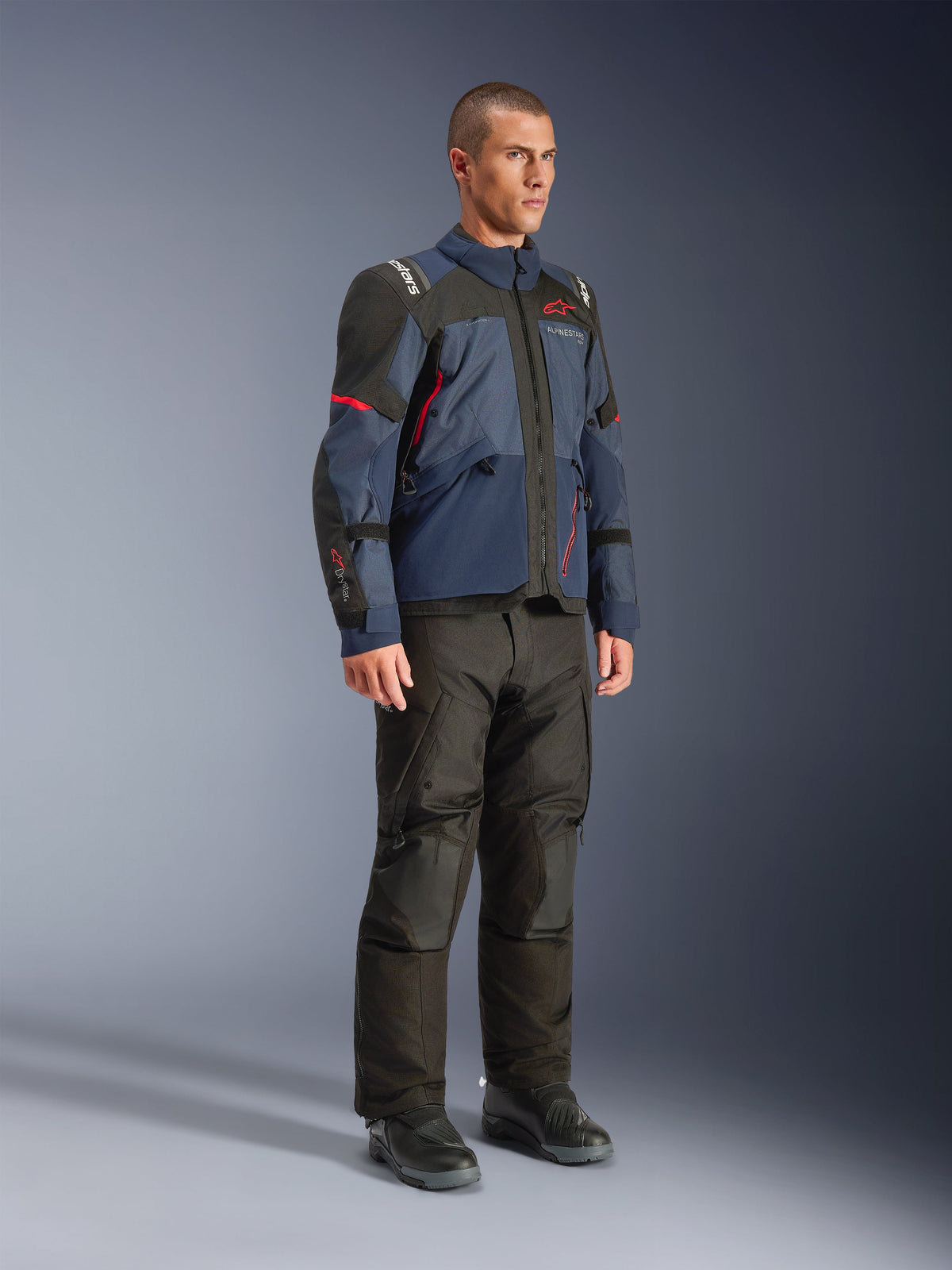 Mandlig model iført Alpinestars Andes V4 Drystar®-jakke i Night Navy og Mood Indigo med sorte paneler og røde detaljer, kombineret med sorte motorcykelbukser og støvler, teknisk adventure touring-udstyr i et studiemiljø.