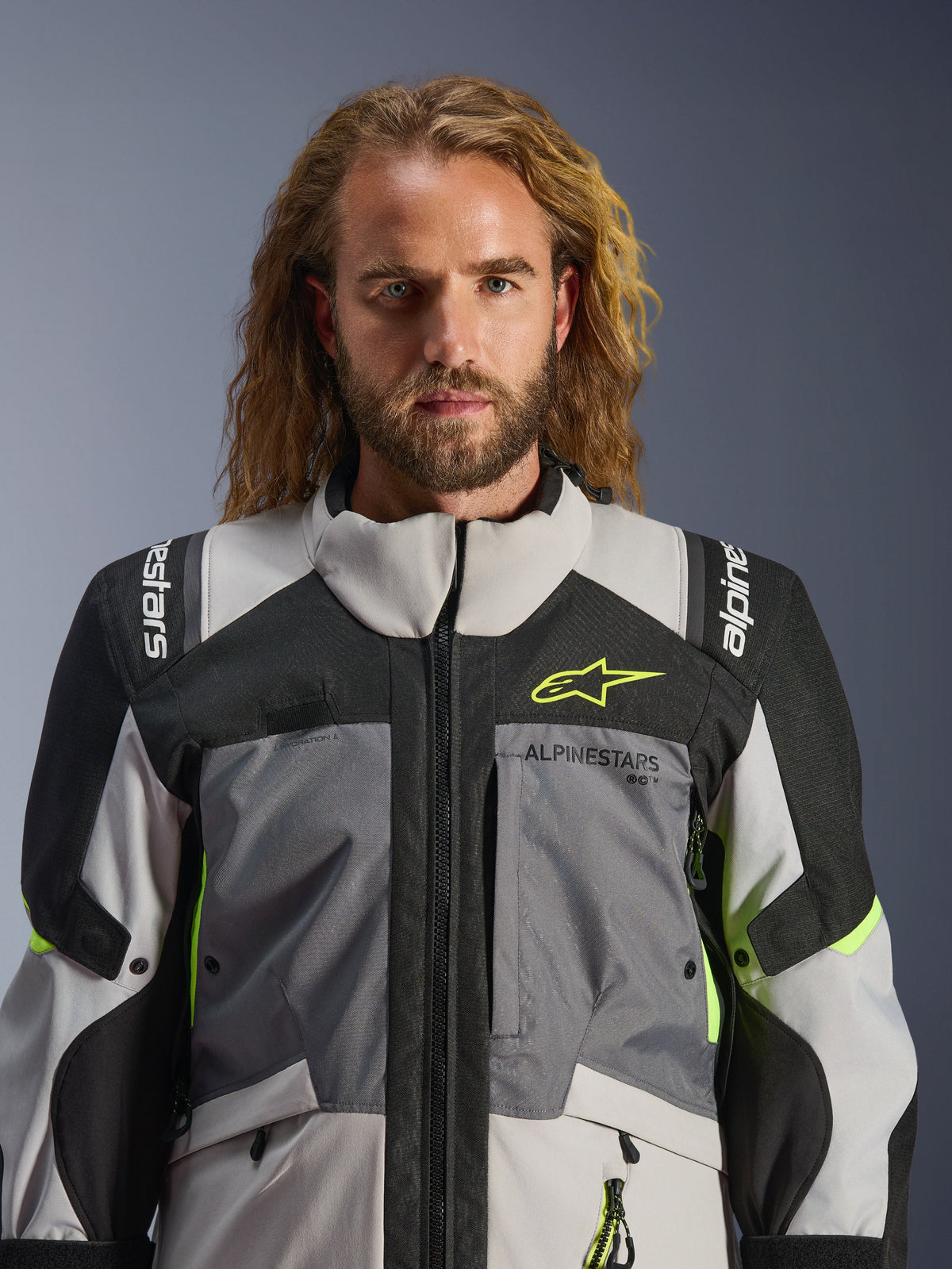 Alpinestars Andes V4 Drystar® Jacket, Dark Gray Ice Gray Black and Yellow Fluo, worn by a man with long wavy hair, featuring technical multi-fabric construction, Alpinestars branding on chest and shoulders, high collar design for adventure riding