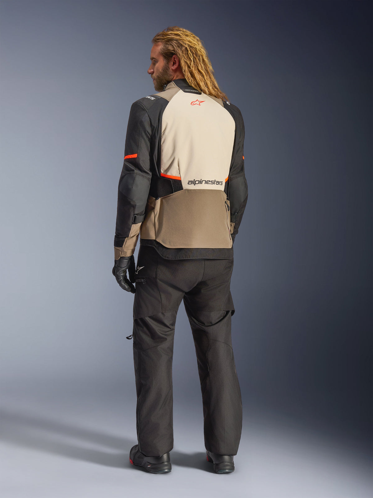 Alpinestars Andes V4 Drystar® jakke, Walnut Mastic Black, set bagfra på mandlig model, med beige og sorte forstærkede tekstilpaneler og orange detaljer, Alpinestars-logo på ryggen, båret med matchende sorte adventure-kørebukser og handsker.