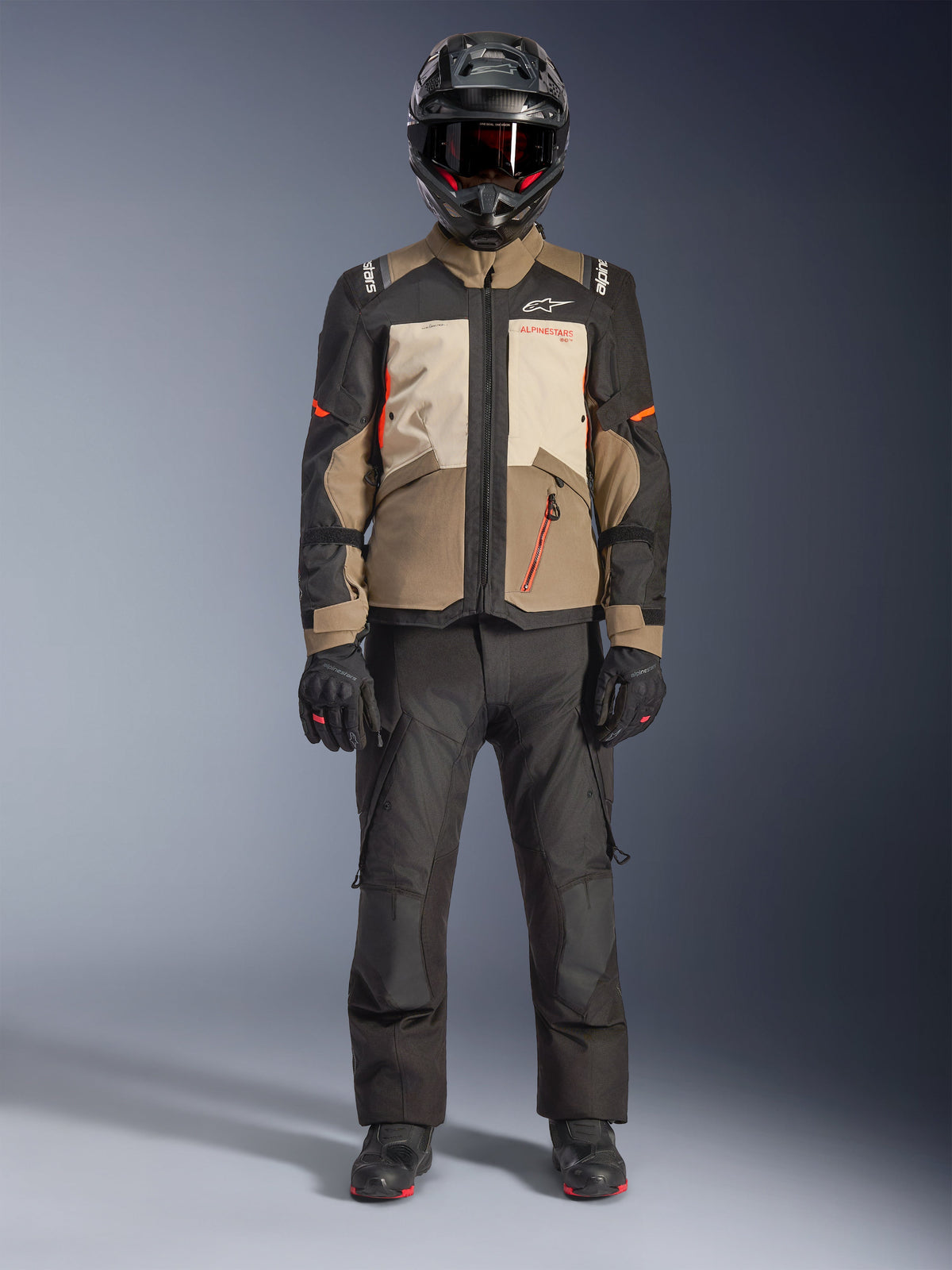 Alpinestars Andes V4 Drystar® Jakke, Walnut Mastic Black, adventure touring motorcykeljakke med tan- og cremefarvede paneler og orange detaljer, båret af en rytter med sort adventure-hjelm, sorte motorcykelbukser og sorte støvler, fuldkropsvisning i studie.