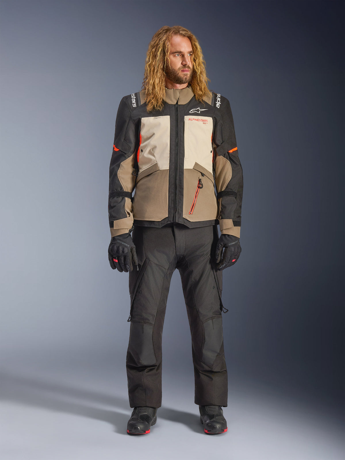 Alpinestars Andes V4 Drystar®-jakke, Walnut Mastic Black, båret med sorte motorcykelbukser og handsker, med et multi-panel adventure-design med orange detaljer og Alpinestars-branding, teknisk tekstiludstyr til kørsel i al slags vejr