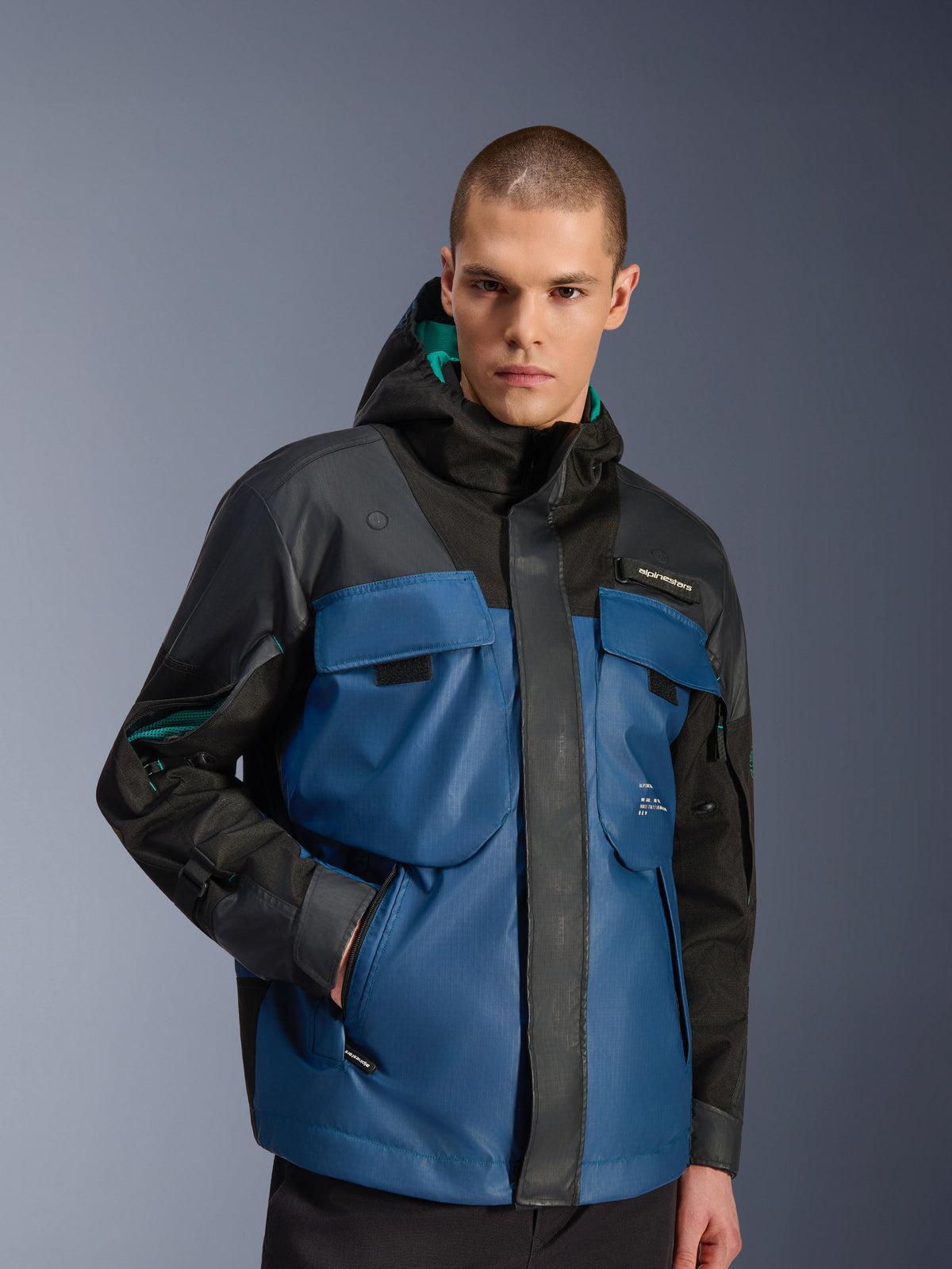 Male model wearing the Alpinestars Mospher Waterproof Jacket in Black and Dark Petrol, featuring large utility chest pockets, a hooded design with teal lining, and technical ripstop fabric for urban motorcycle commuting.