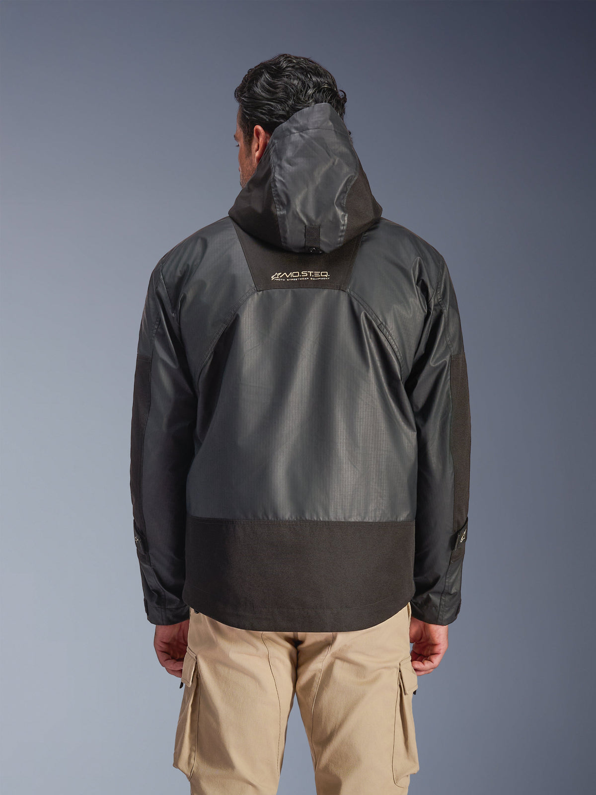 Rear view of a man wearing the Alpinestars Mospher Waterproof Jacket in Black, featuring a hood and durable ripstop textile construction, shown with tan cargo pants, highlighting the MO.ST.EQ. logo on the upper back, urban motorcycle gear design.