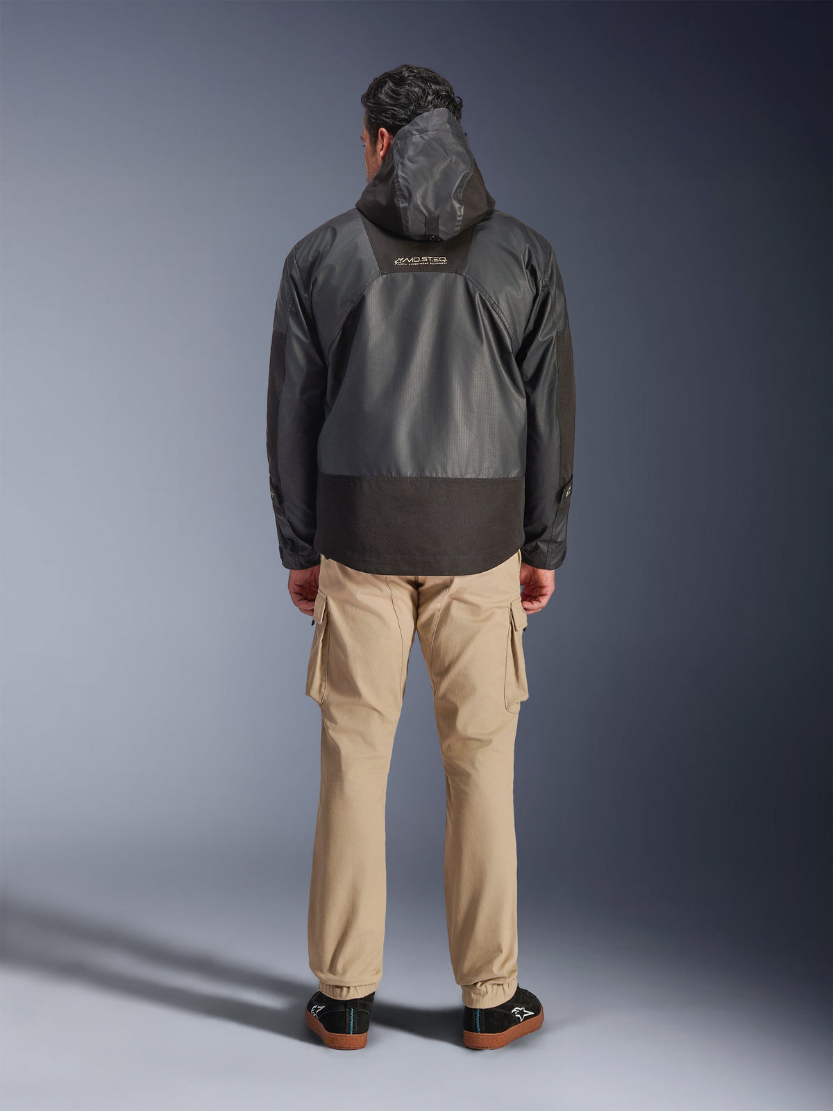 Alpinestars Mospher Waterproof Jacket, Black motorcycle parka shown from the back, featuring MO.ST.EQ logo on upper back and ripstop fabric construction, worn with tan cargo pants and black sneakers, urban commuter style.