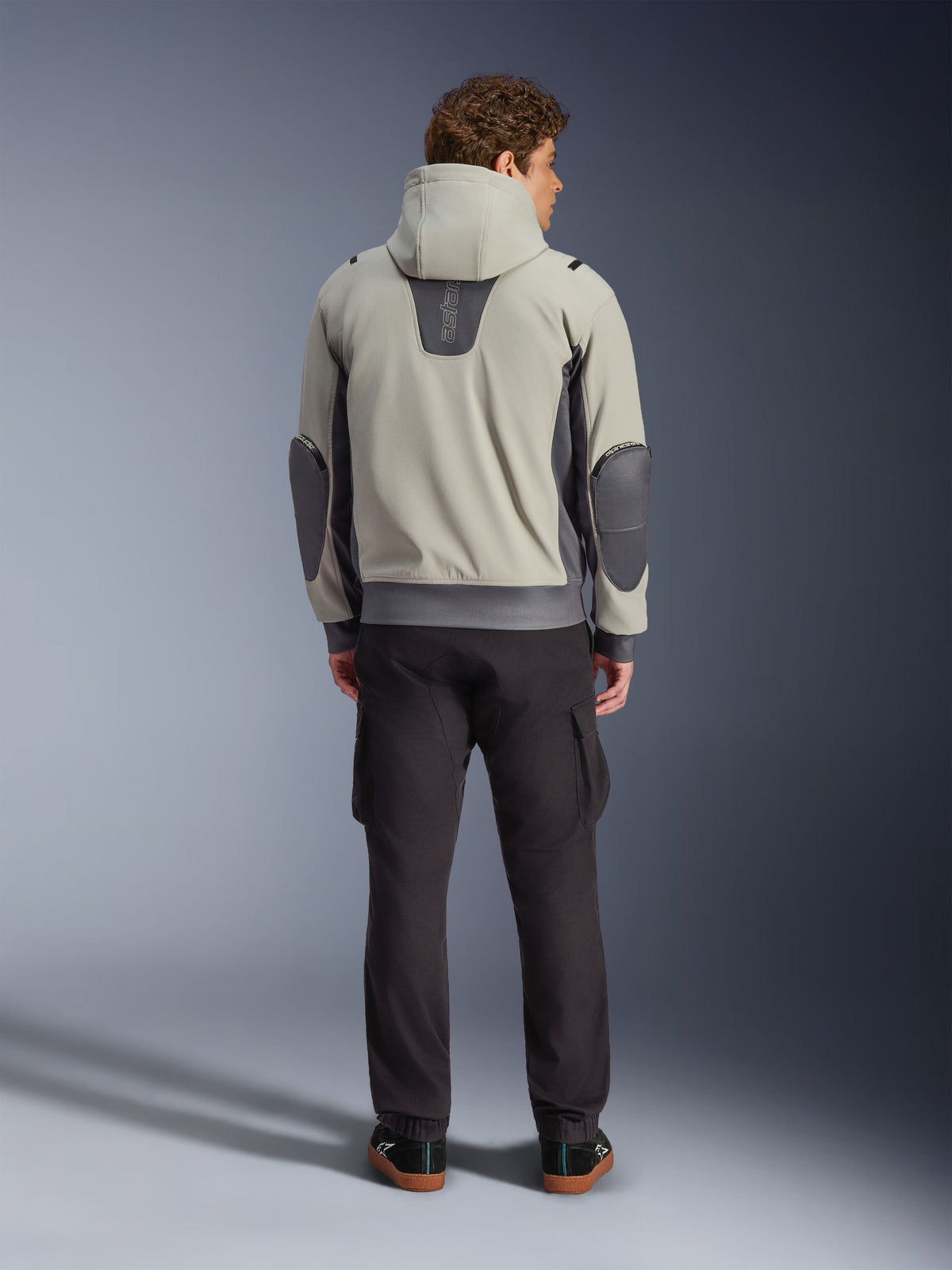 Alpinestars Moblast Waterproof Jacket, Iron Gray Dark Sage, set bagfra af en mandlig kører iført motorcykeljakke med hætte, lysegrå og mørk salviegrøn farvekombination, lodret 'astars' logo på den øverste del af ryggen, mørkegrå albuelapper, stylet med mørke cargo-bukser for et urbant, moderne look