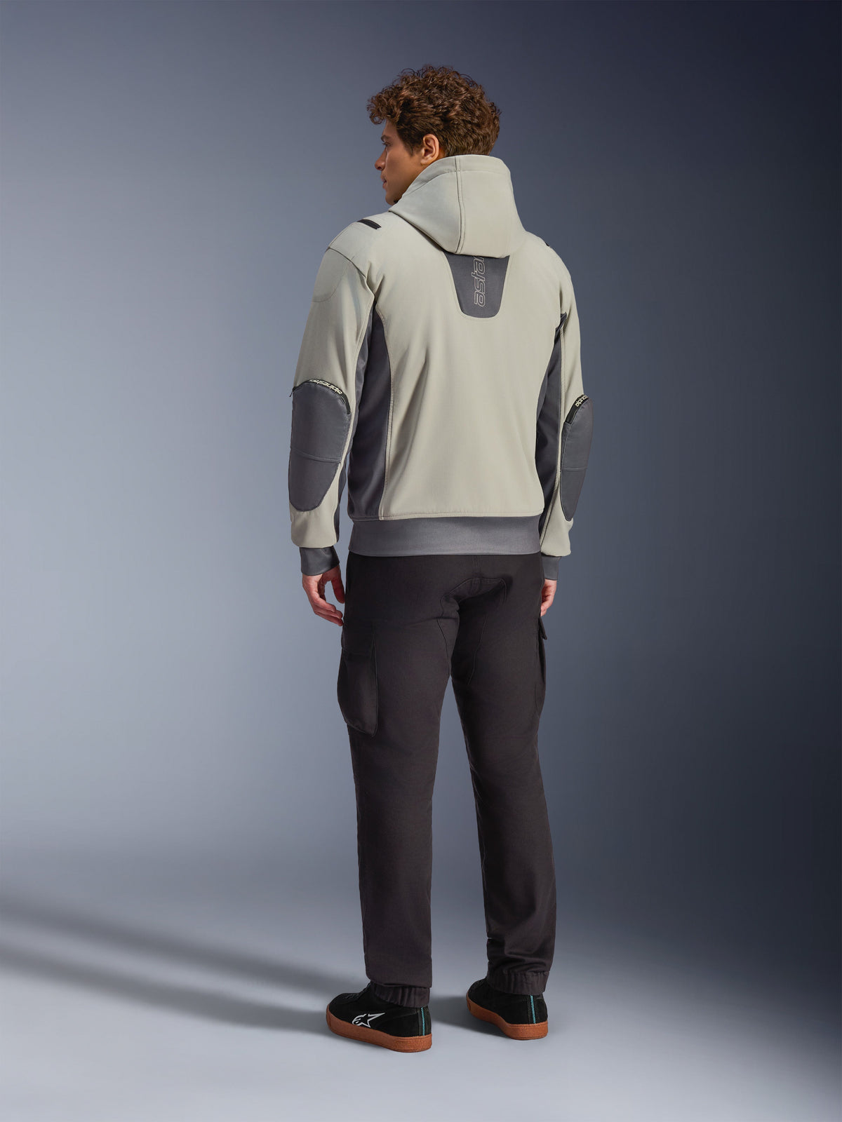 Mandlig model set bagfra iført Alpinestars Moblast Waterproof Jacket i Iron Gray Dark Sage, urban motorcykeljakke med hætte, mørkegrå sidepaneler og forstærkede albuelapper, kombineret med mørke kørebukser og sorte sneakers, moderne minimalistisk æstetik til urban pendling