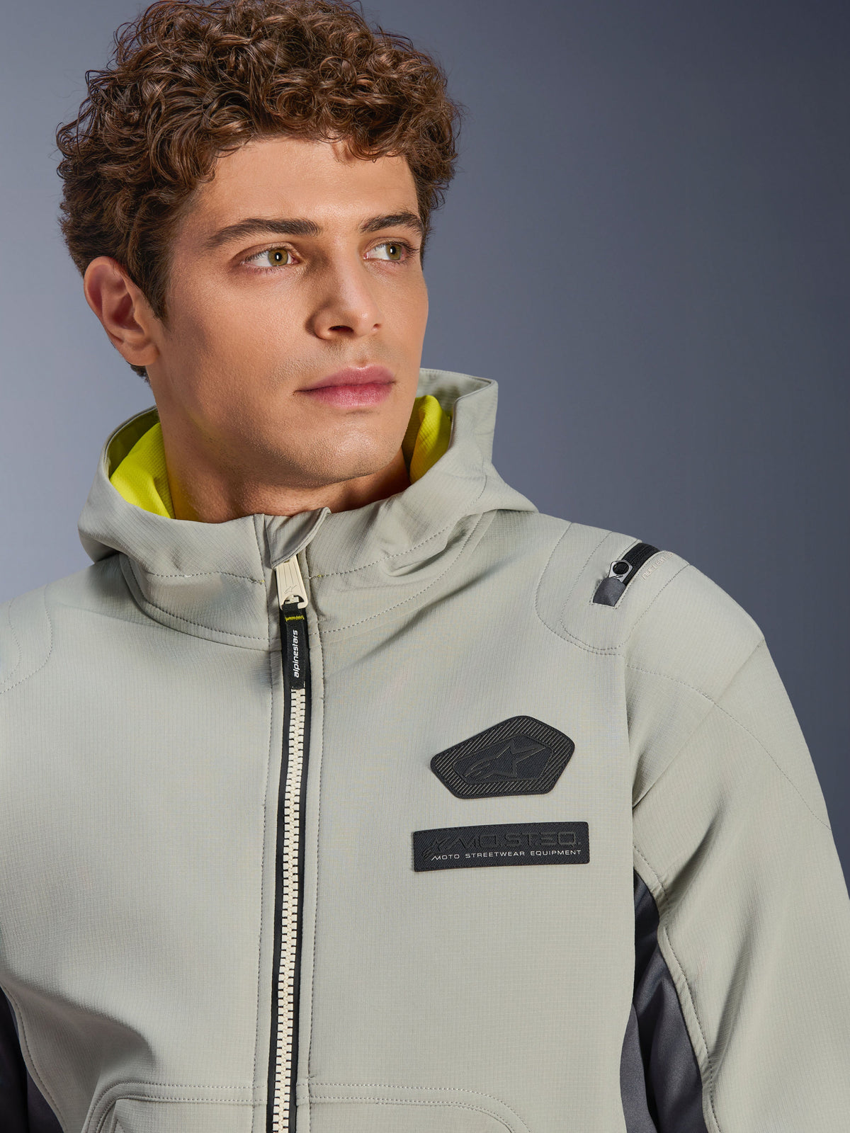 Alpinestars Moblast vandtæt jakke, Iron Gray Dark Sage, nærbillede af mandlig model iført jakken, med sort Alpinestars-logo og MO.ST.EQ. brystmærker, lysegult hættefor og et moderne teknisk design.