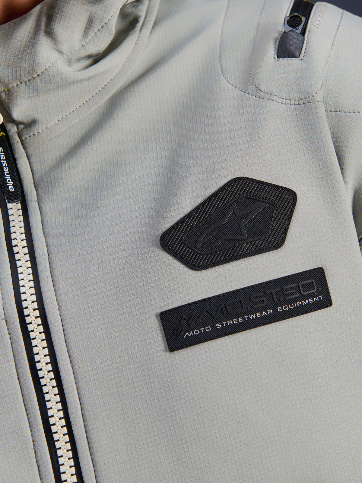 Alpinestars Moblast vandtæt jakke, Iron Gray, nærbillede af brystmærker med Alpinestars-logo og MO.ST.EQ. Moto Streetwear Equipment-branding, teknisk ripstop-materiale med kraftig lynlås foran og detalje på skulderlomme