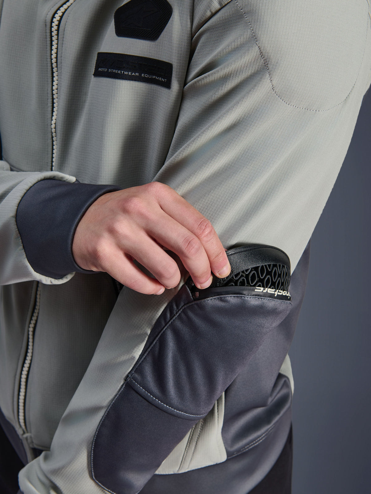 Alpinestars Moblast vandtæt jakke, Iron Gray og Dark Sage, nærbillede der viser fjernelse af Nucleon Flex Plus-beskyttelse fra den udvendige lynlåslomme på albuen, teknisk tekstil urban motorcykelbeklædning med MO.ST.EQ.-branding