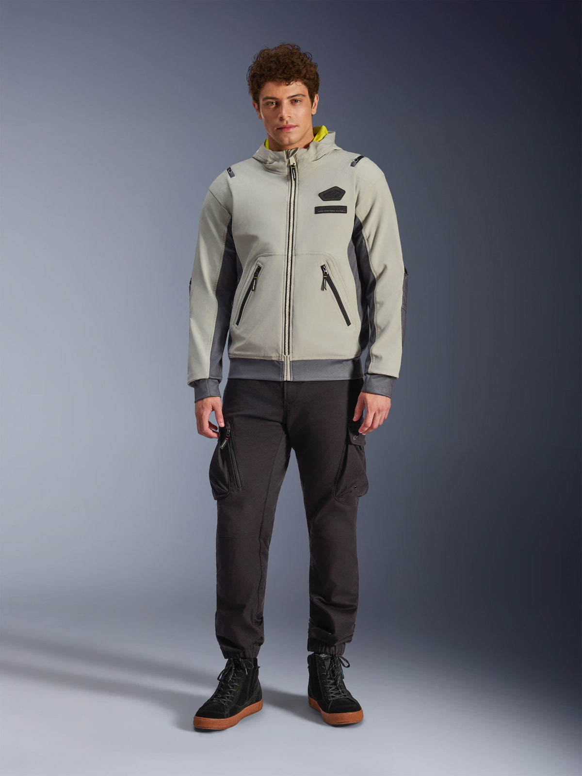 Mandlig model iført Alpinestars Moblast Waterproof Jacket i Iron Gray og Dark Sage, motorcykeljakke med hætte og et moderne teknisk design, kombineret med mørke cargo-kørebukser og sorte støvler, urban moderne motorsportbeklædning