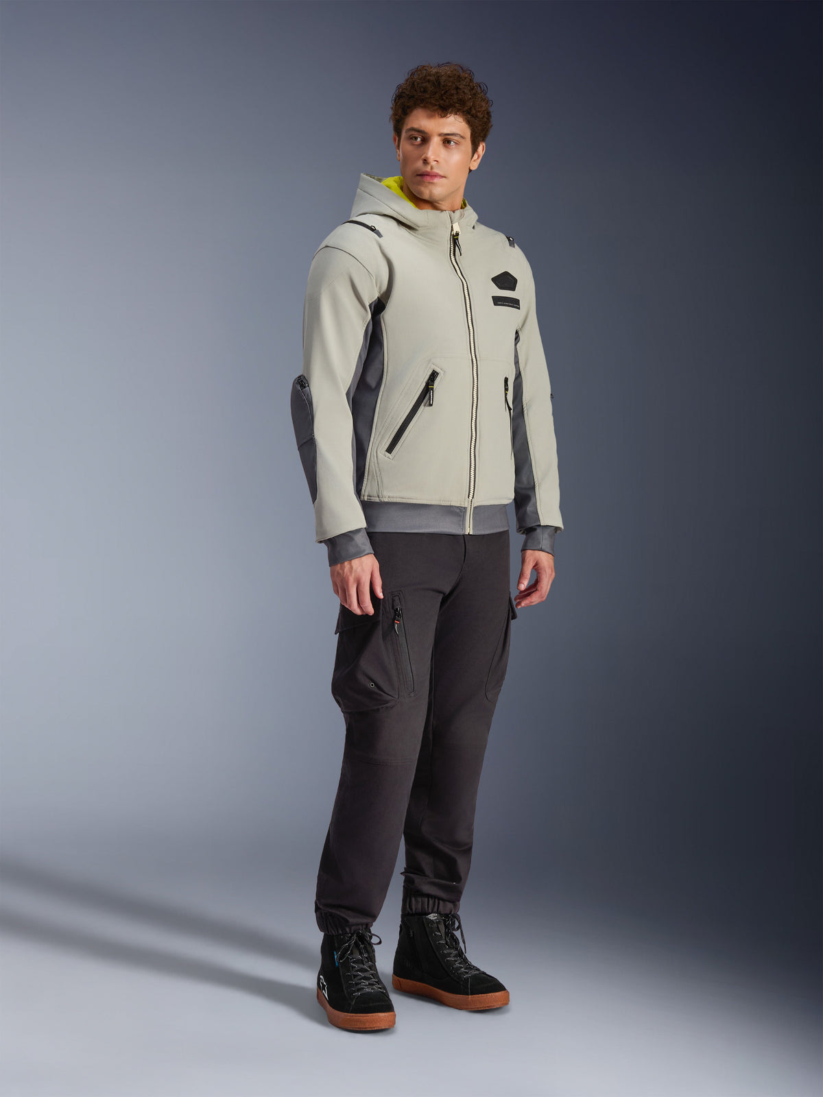 Alpinestars Moblast vandtæt jakke, Iron Gray og Dark Sage, urban motorcykeljakke med hættetrøjedesign, båret af en model med mørke kørebukser og sorte støvler, minimalistisk cyber-tech-æstetik med gult indvendigt for i hætten og vandtætte lynlåse