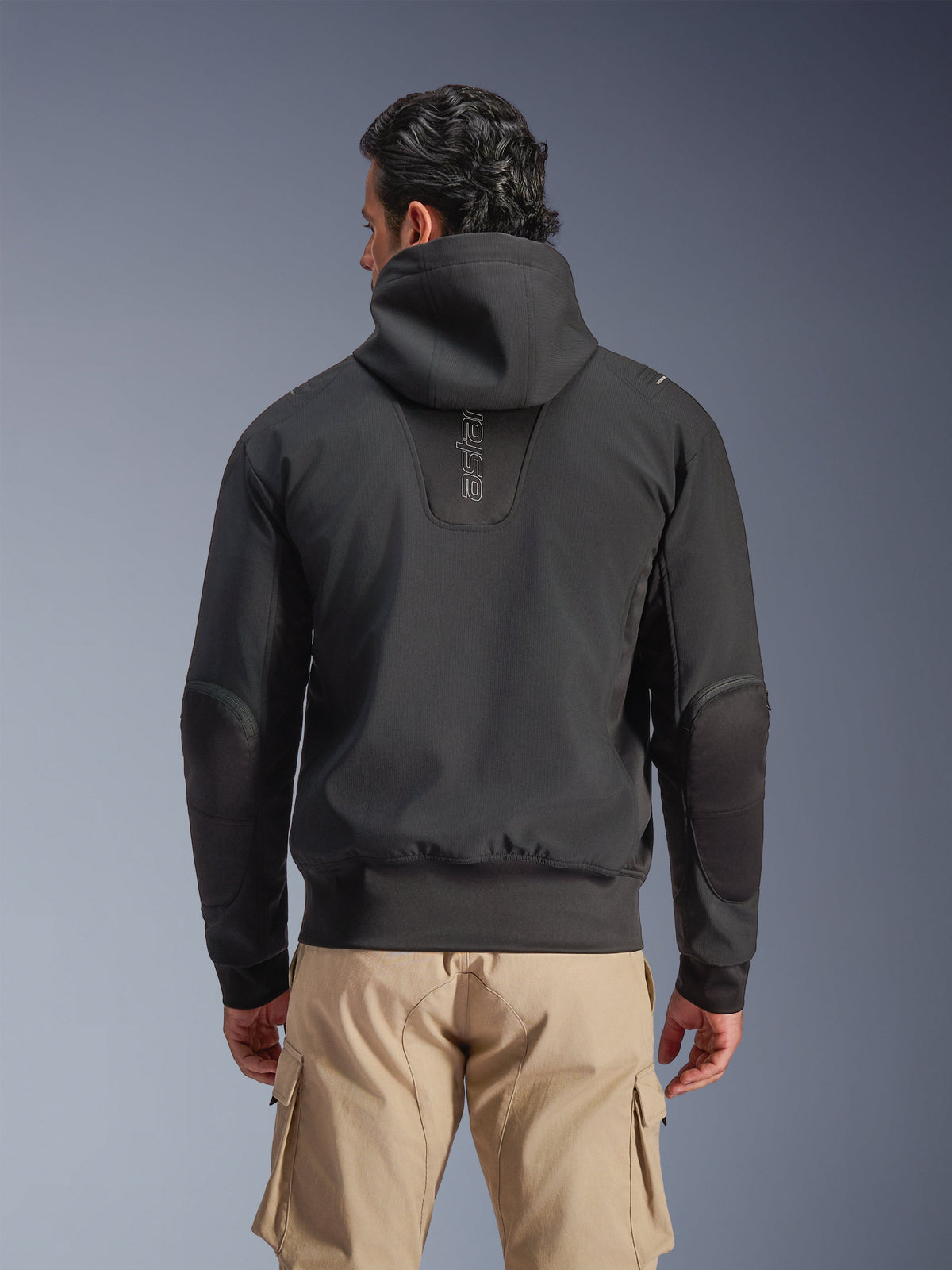 Alpinestars Moblast vandtæt jakke, sort, bagfra af urban motorcykeljakke med hætte, med lodret 'astars' logo på ryggen, elastik i talje og manchetter, forstærkede albuer og moderne teknologisk design, båret med sandfarvede cargo-bukser.