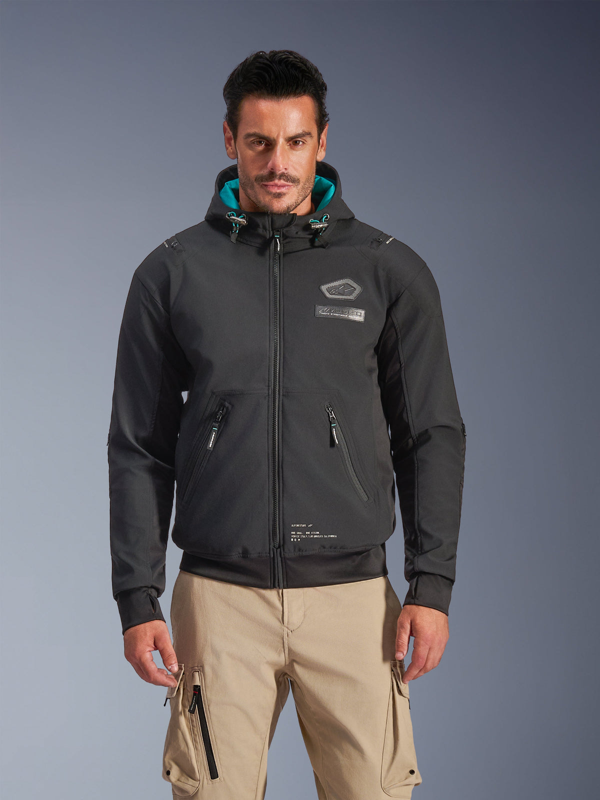 Alpinestars Moblast Waterproof Jacket, urban motorcykeljakke til mænd, sort med turkis hættefor, 3D-lynlåslommer og design med hætte, båret med sandfarvede cargobukser, moderne urbant motorcykeltøj