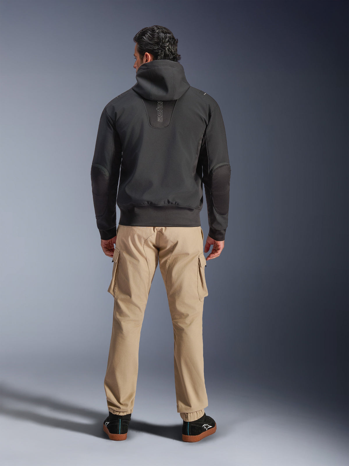 Alpinestars Moblast vandtæt jakke, sort, urban motorcykel-hættetrøje set bagfra, båret med sandfarvede cargobukser og sorte sneakers, minimalistisk design med lodret 'astars'-branding på den øverste del af ryggen, moderne køreudstyr til al slags vejr