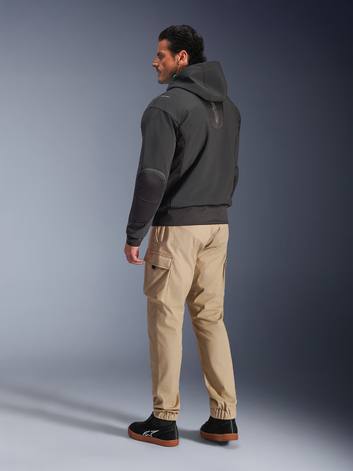 Alpinestars Moblast Vandtæt Jakke, sort motorcykel tech-hættetrøje, set fra siden og bagfra, båret med beige cargobukser og sorte sneakers, design med hætte, teknisk tekstilkonstruktion og moderne urban æstetik