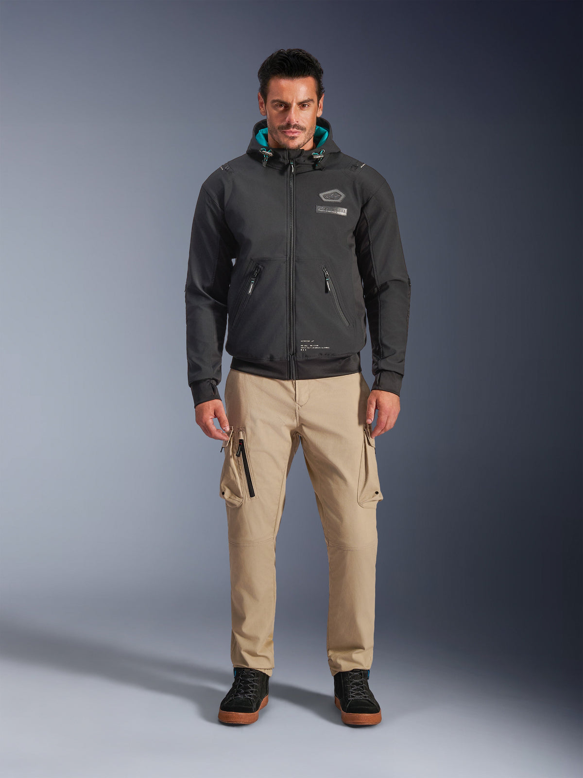 Alpinestars Moblast Waterproof Jacket, sort urban motorcykel-hættetrøje med blågrønt indvendigt hættefor, båret med sandfarvede cargobukser og sorte high-top støvler, moderne køreudstyr til alle årstider