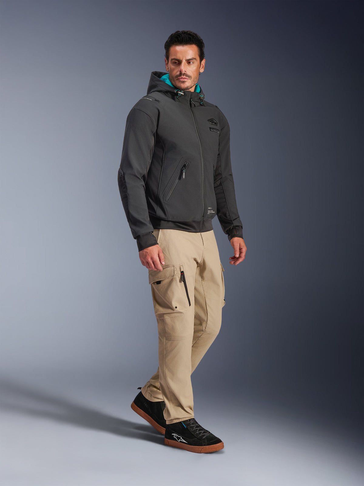 Mandlig model iført Alpinestars Moblast Waterproof Jacket i sort, urban motorcykel-hættetrøje med teal-farvet for i hætten, kombineret med khaki cargobukser og sorte sneakers, moderne minimalistisk design med teknisk strækstof til pendling i al slags vejr